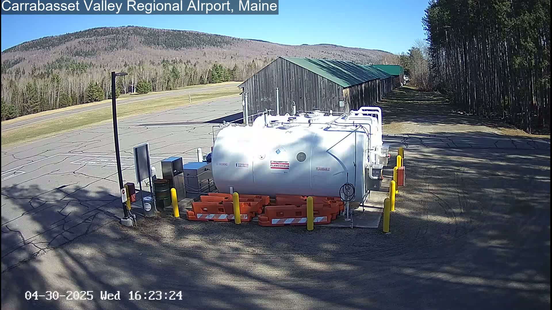 A large white tank sits in a paved area of an airport on a sunny day, near a long wooden building and a stand of pine trees.
