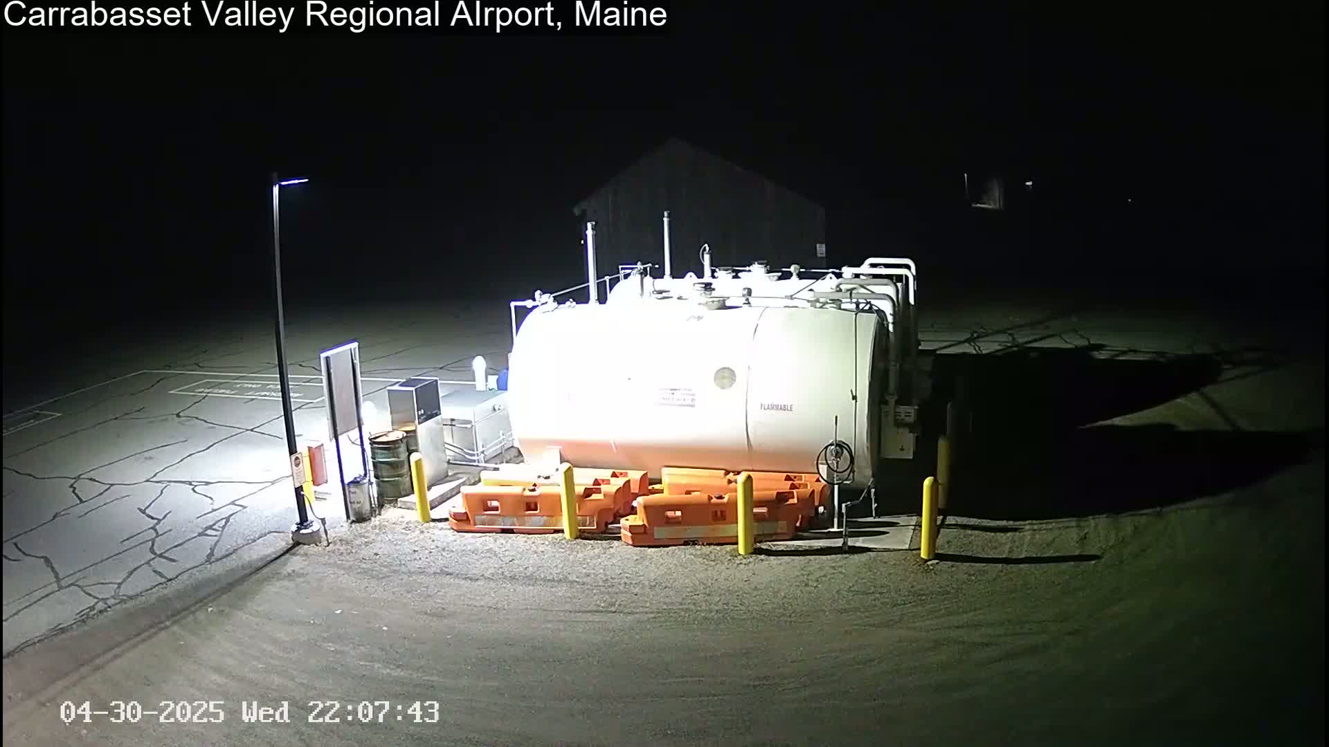 At night, a large white fuel tank sits in a dark, cracked asphalt lot, illuminated by a single lamppost.