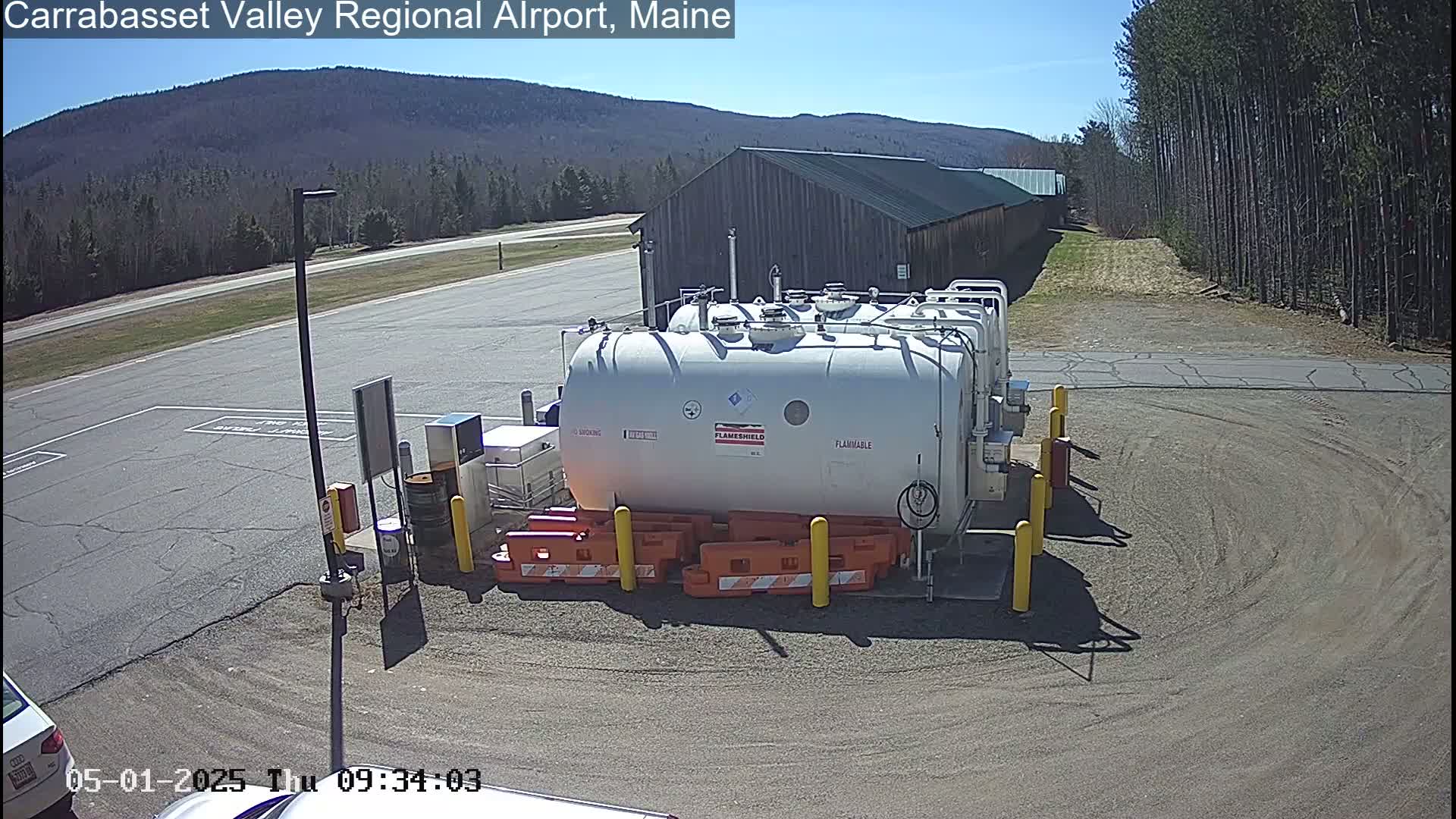 A large white fuel tank sits in a parking lot on a sunny day, with a long building and forested hills in the background.