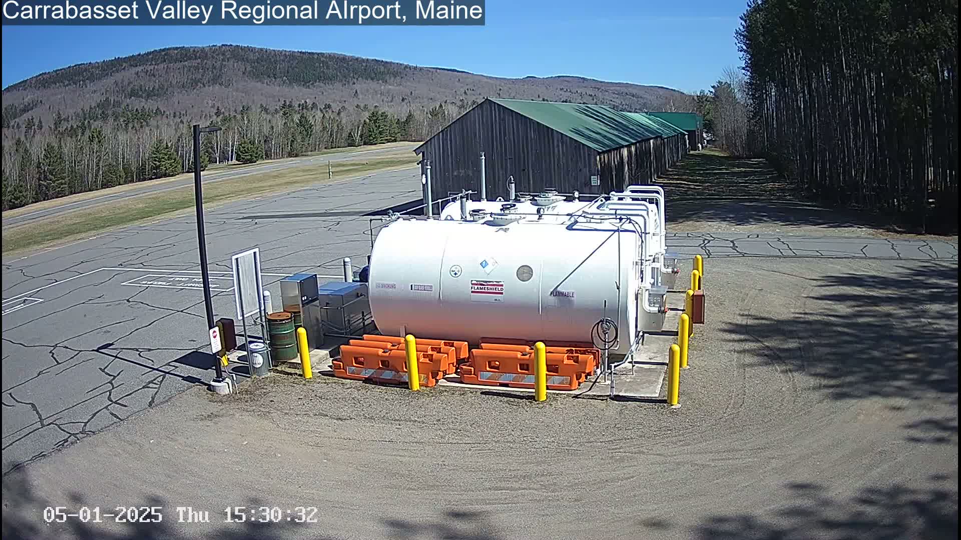 A large white fuel tank sits in a paved area next to a long barn-like building, with trees and mountains visible in the background under a clear blue sky.