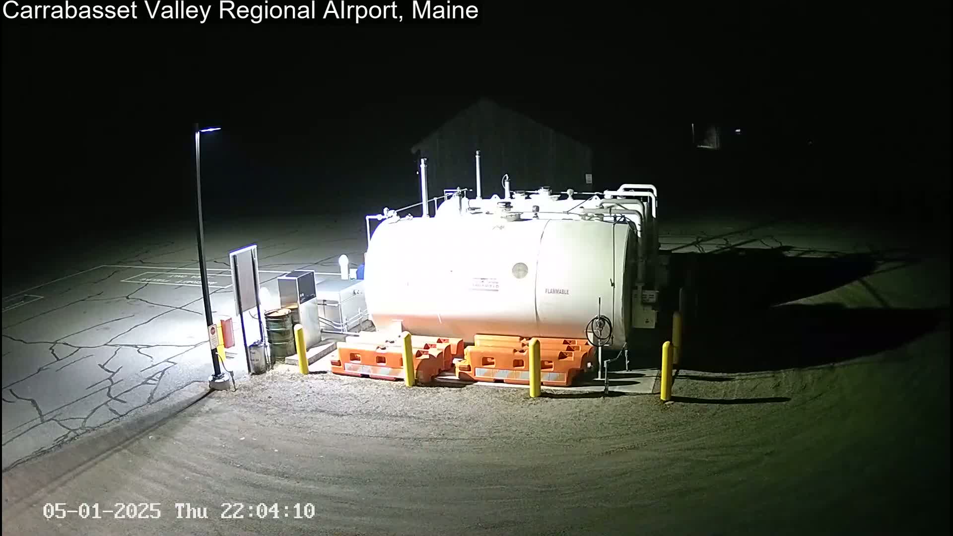 A large white fuel tank sits in a dark, nighttime airport parking lot illuminated by a single lamppost.
