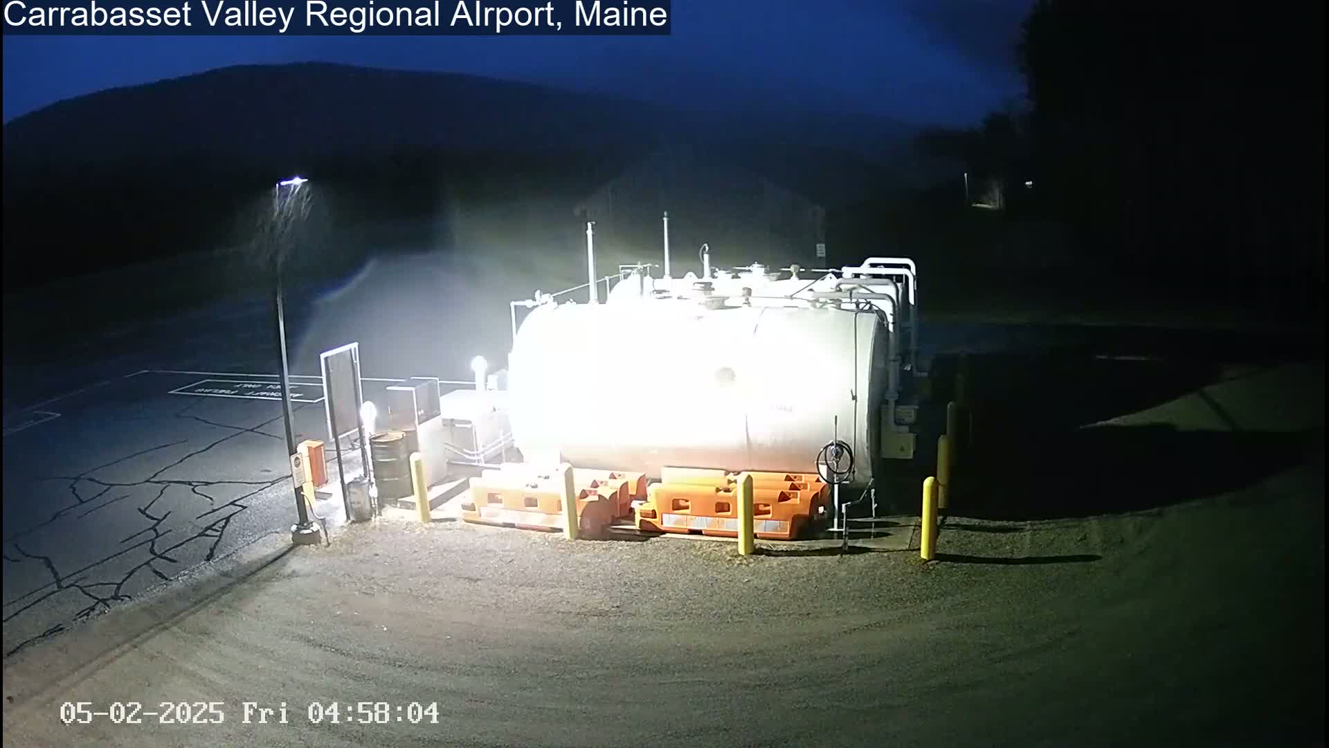 A large, cylindrical storage tank sits in a dimly lit, rain-swept asphalt lot at night.