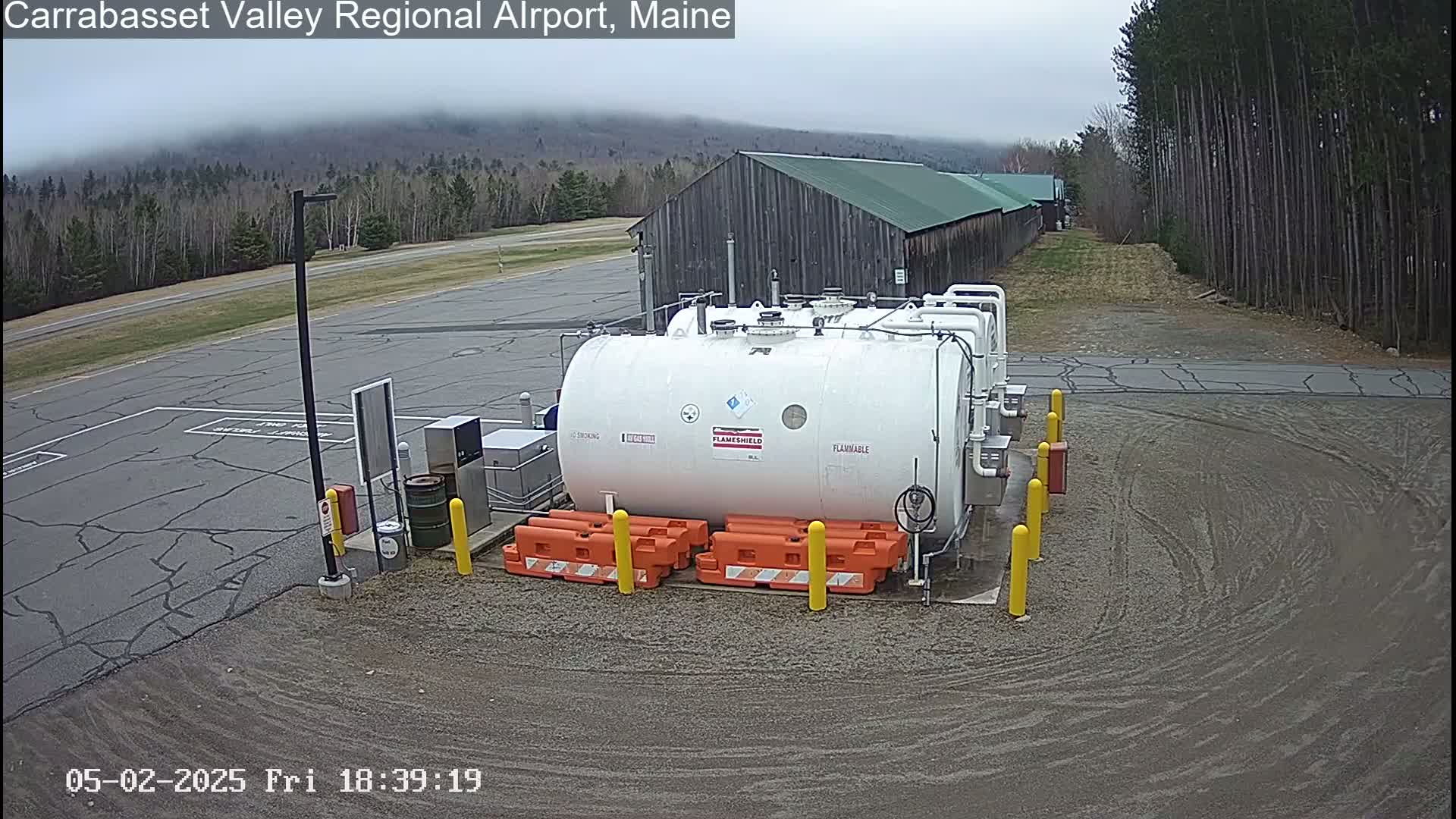 A large white cylindrical tank labeled "flammable" sits in a gravel area next to a weathered wooden building under an overcast sky, surrounded by trees and pavement.