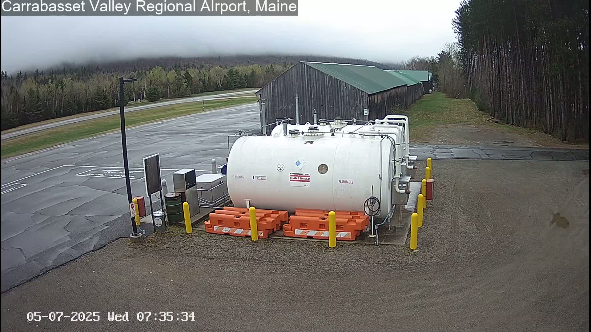 A large white fuel tank sits in a gravel lot next to a long, low wooden building on an overcast day, surrounded by trees.