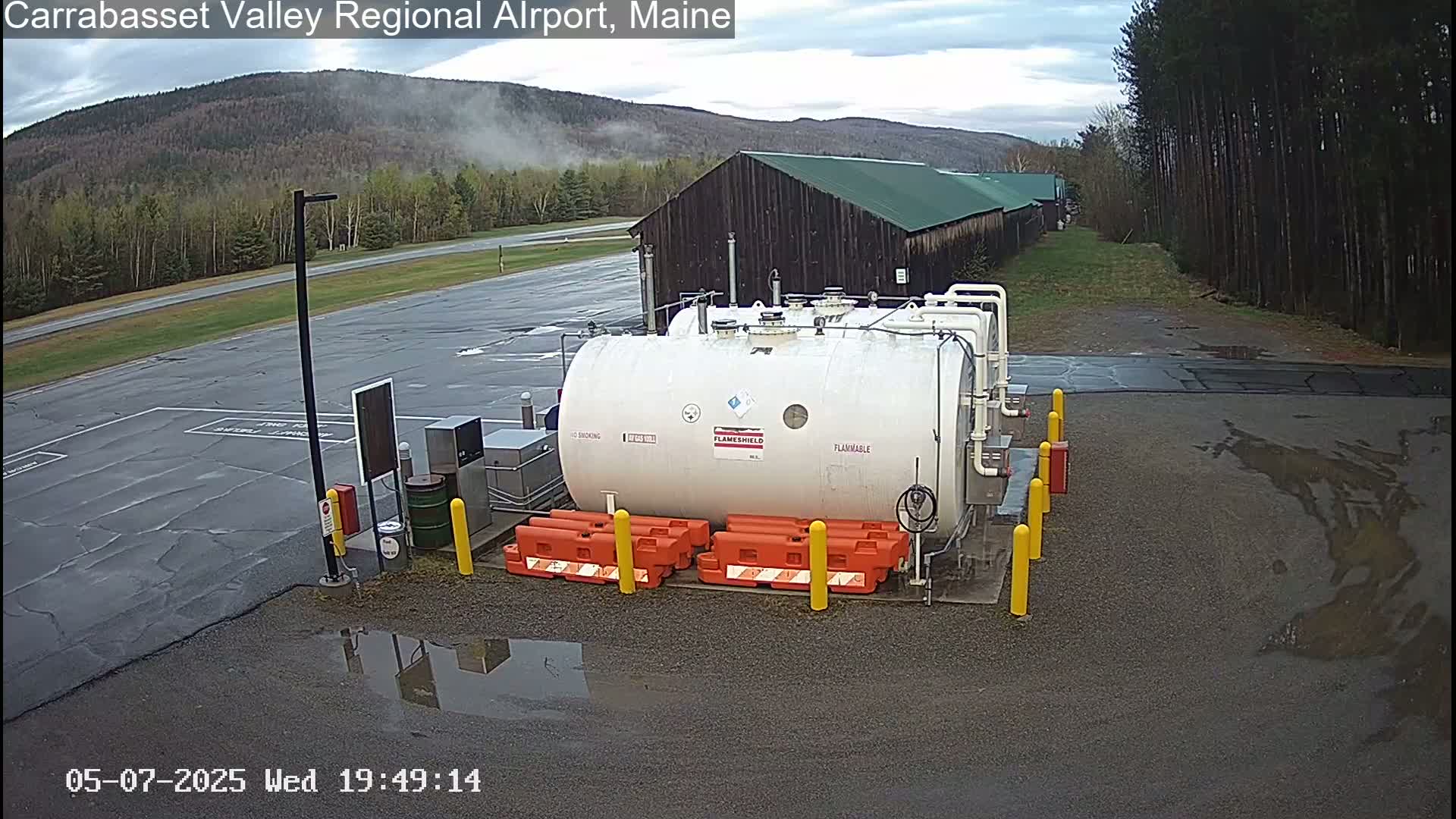 A large white cylindrical tank with the word "Flammable" on it sits in a paved area near a dark-colored building, with trees and a hazy mountain in the background under an overcast sky.