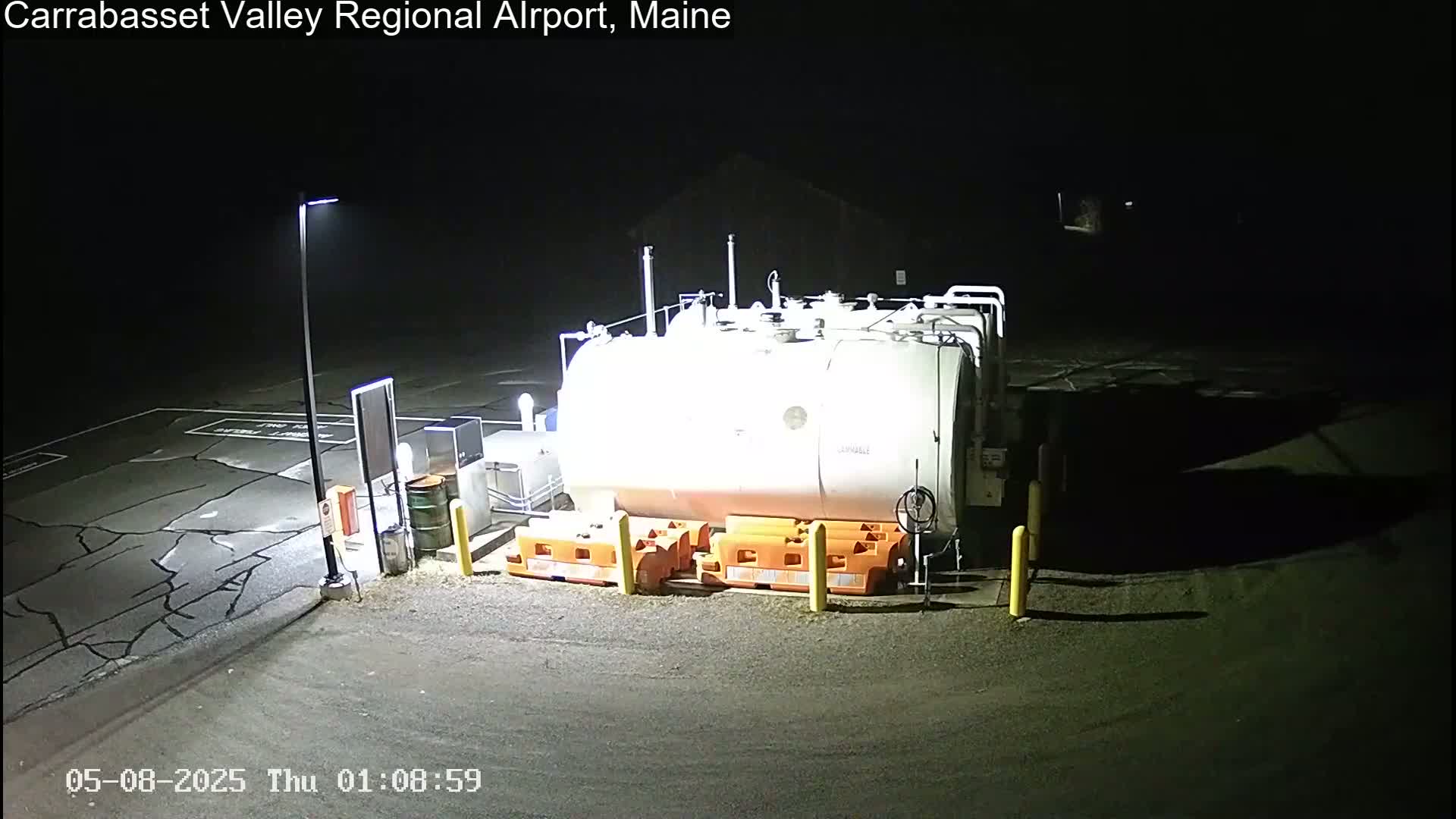 At night, a large white fuel tank sits in a cracked asphalt lot illuminated by a single lamppost, surrounded by orange safety barriers.