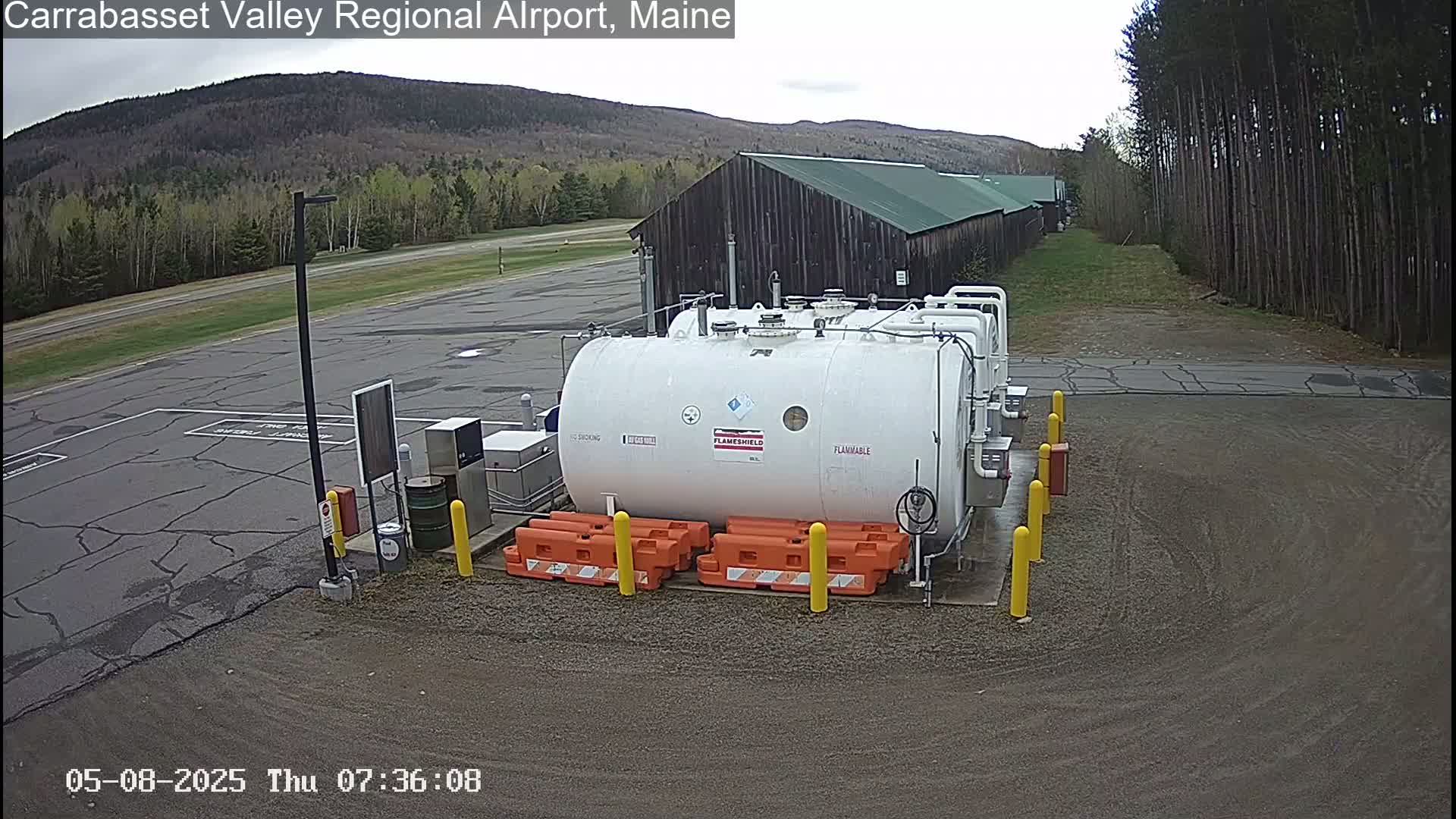 A large white fuel tank sits in a gravel area of an airport, with a dark-colored building and a line of trees in the background on an overcast day.