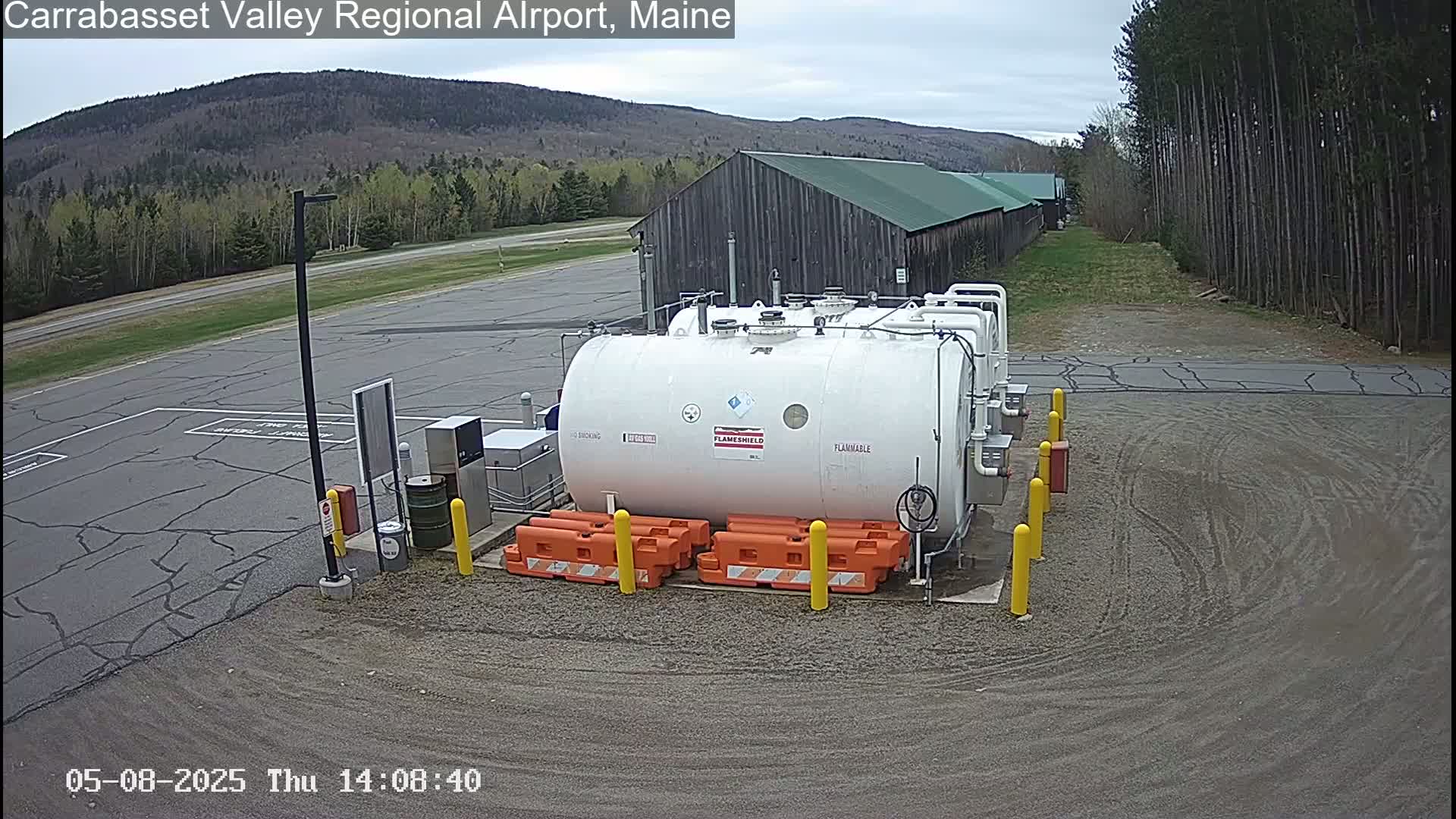 A large white fuel tank sits in a gravel area of an airport, surrounded by orange safety barriers, under a cloudy sky with trees and a mountain in the background.