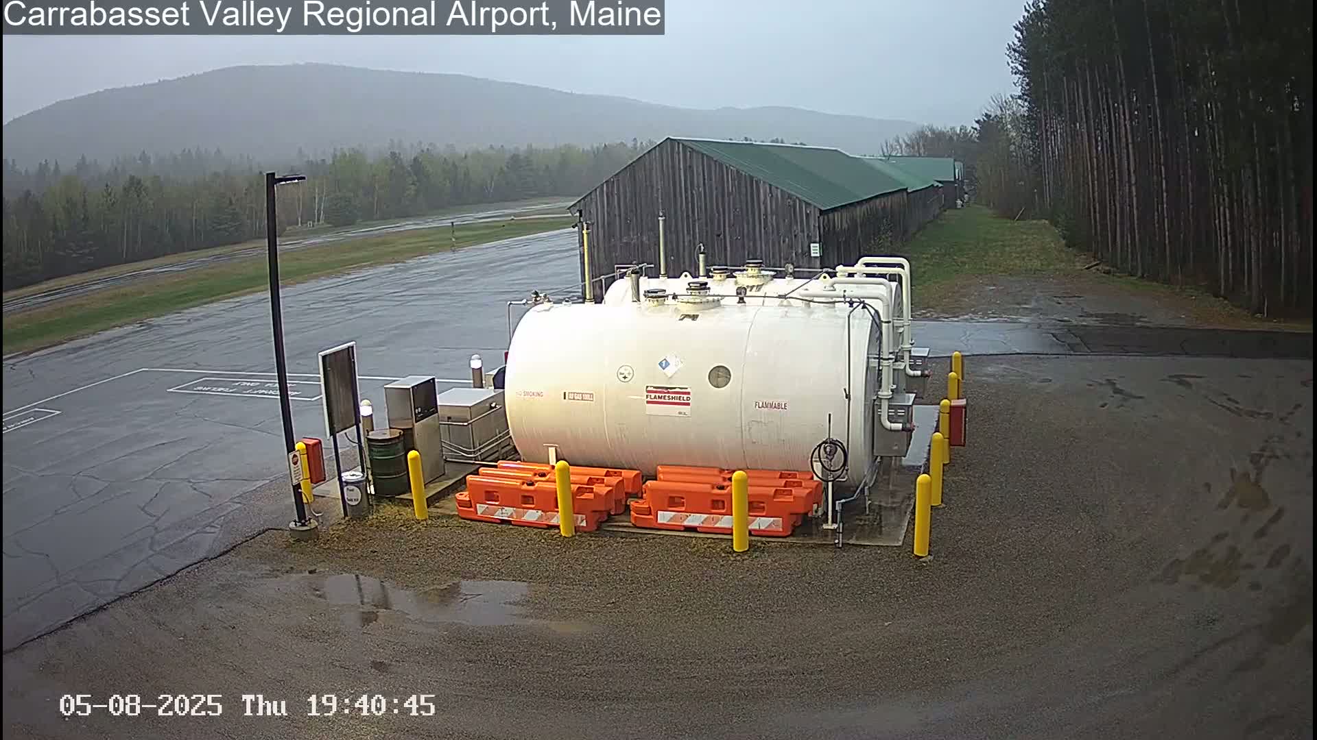 A large white cylindrical fuel tank sits in a wet, outdoor airport area next to a long wooden building under a gray sky.