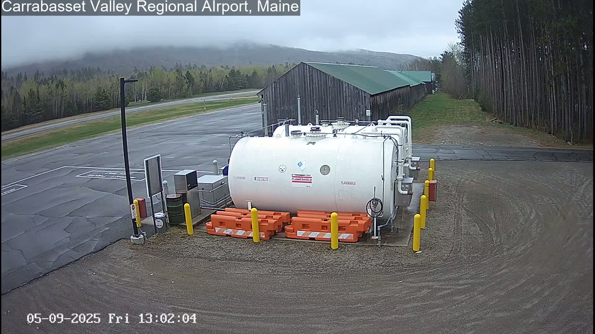 A large white fuel tank sits in a gravel area of an airport on an overcast day, surrounded by orange barriers, with a wood building and trees in the background.