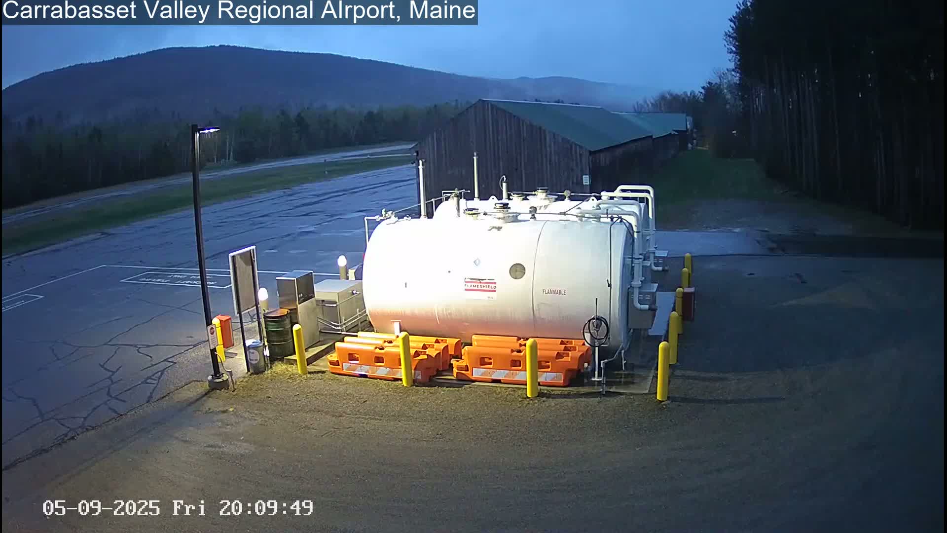 A large white fuel tank sits in a dark, wet airport lot at night, backed by a dark-colored building and trees under a twilight sky.