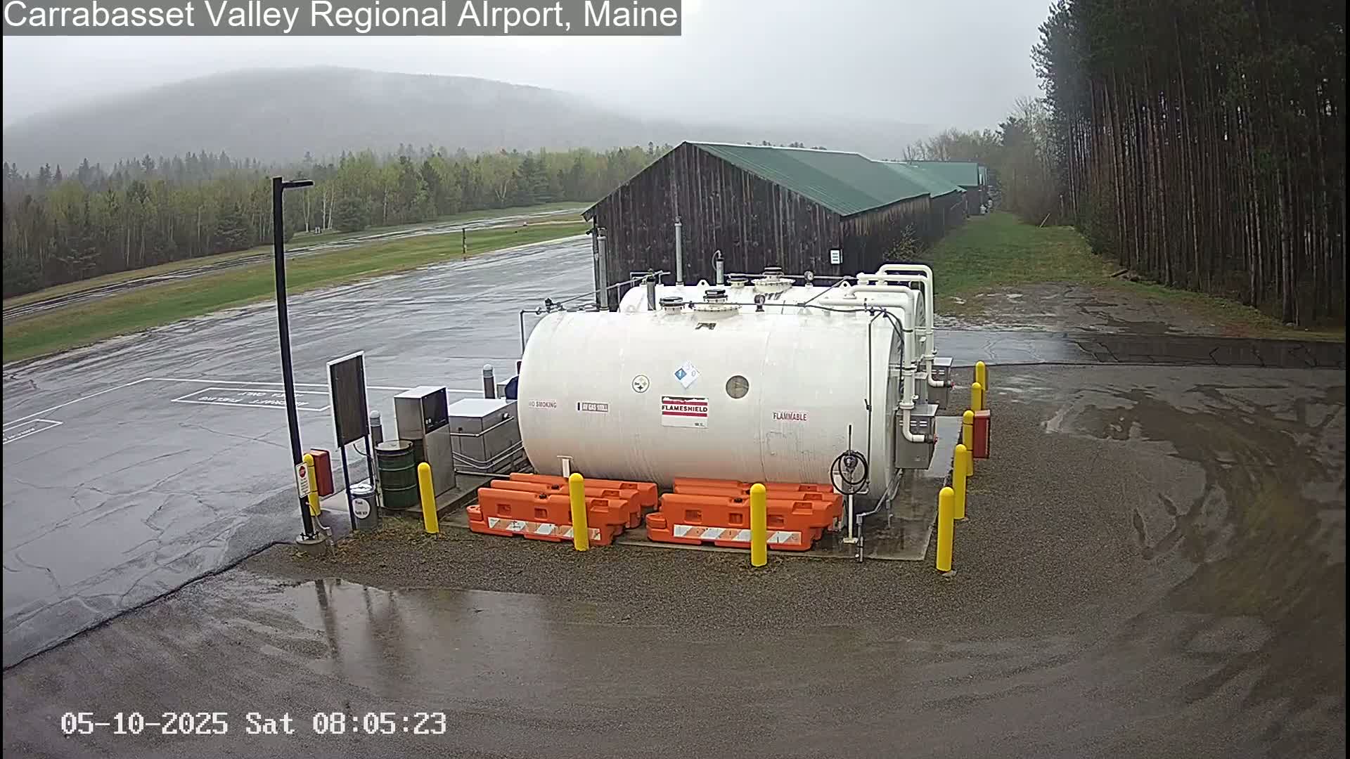 A large white flammable liquid storage tank sits in a wet, empty airport parking lot next to a dark brown building, under an overcast sky.