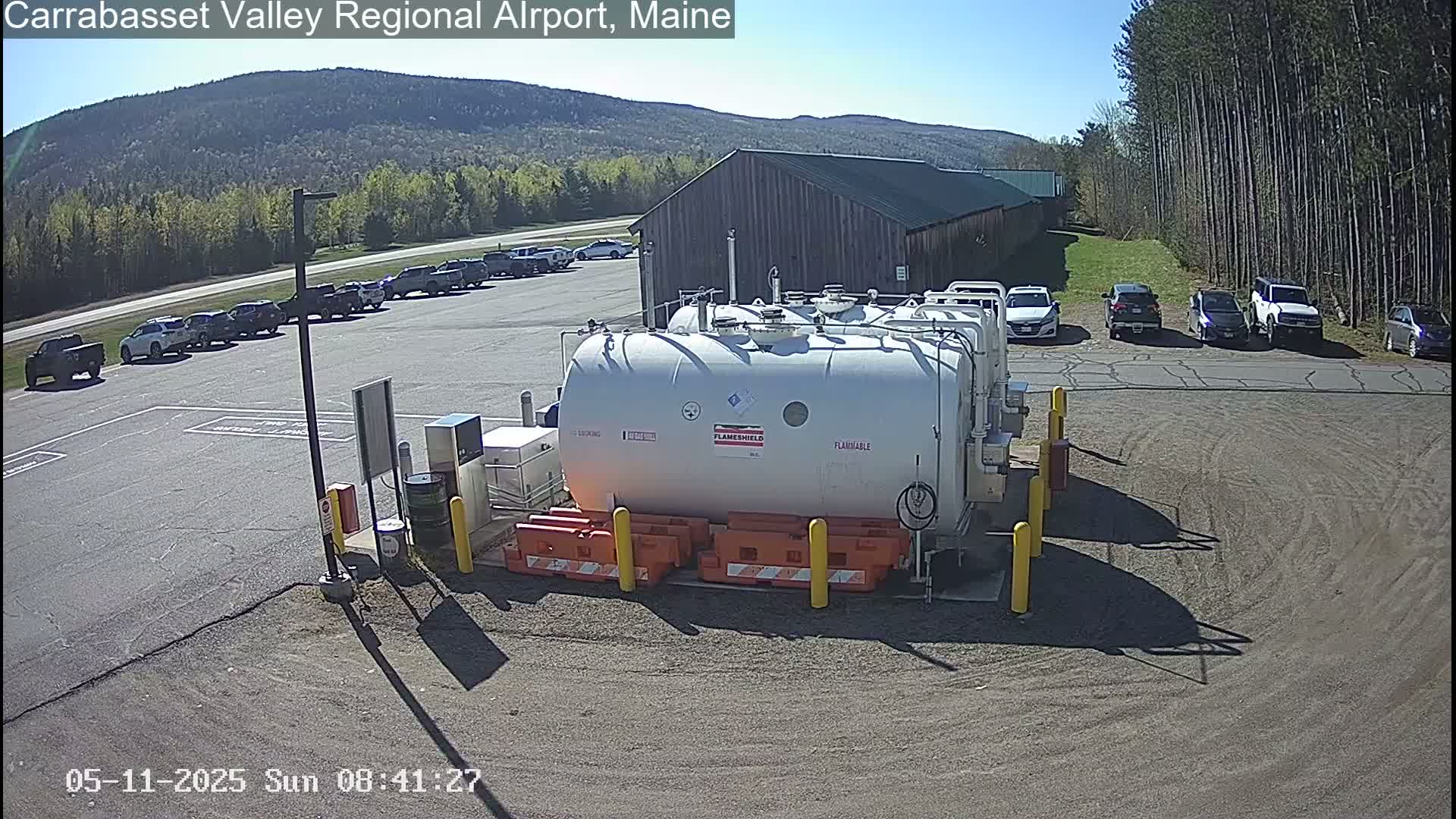 A large white fuel tank sits in a parking lot next to a wood-sided building under a sunny sky, with cars parked nearby and trees and mountains in the background.