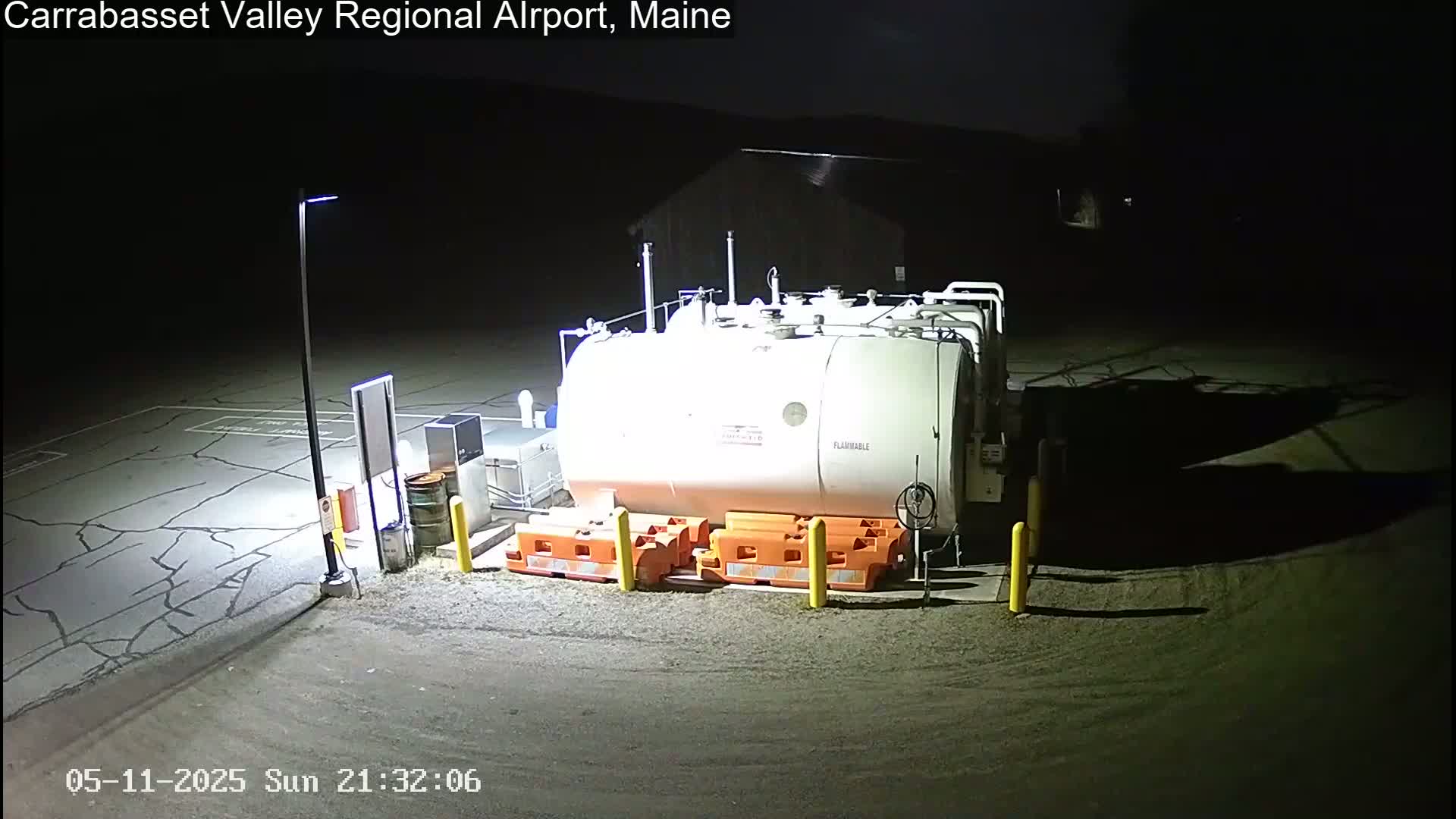 A large, white fuel tank sits in a dimly lit, nighttime airport lot, surrounded by orange and yellow safety barriers.