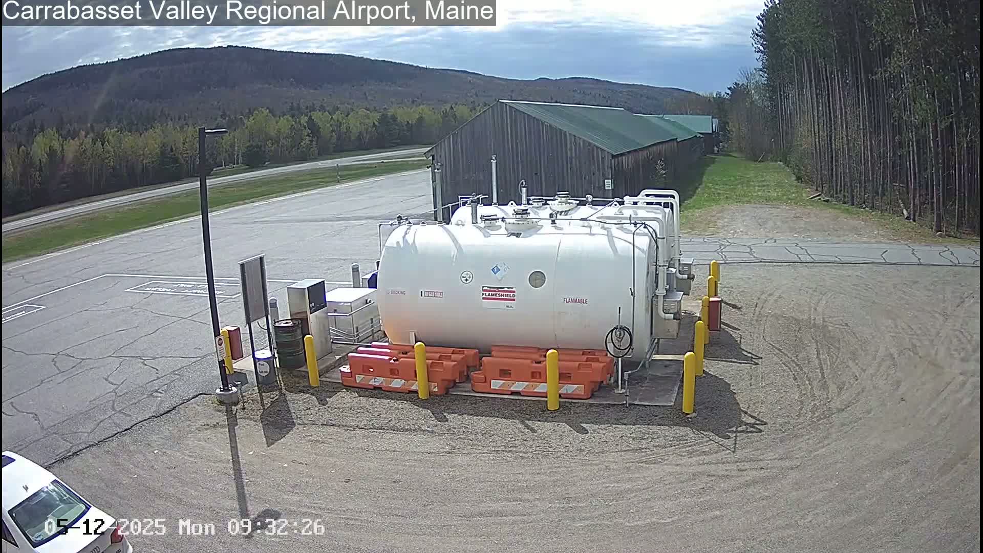 A large white flammable liquid storage tank sits in a paved area of an airport on a partly sunny day, with a wood-sided building and trees in the background.