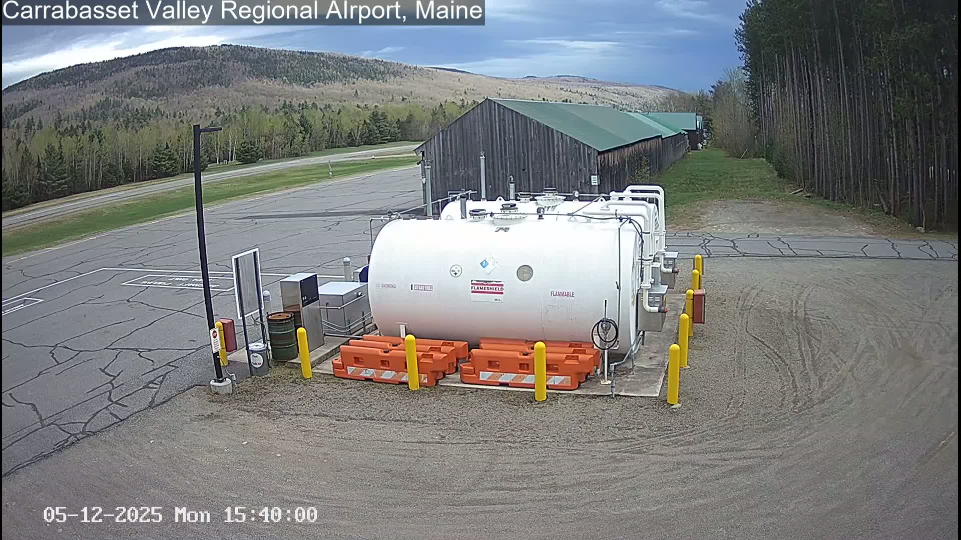 A large white fuel tank sits in a gravel area of an airport, with a barn and a treeline visible in the background under a partly cloudy sky.