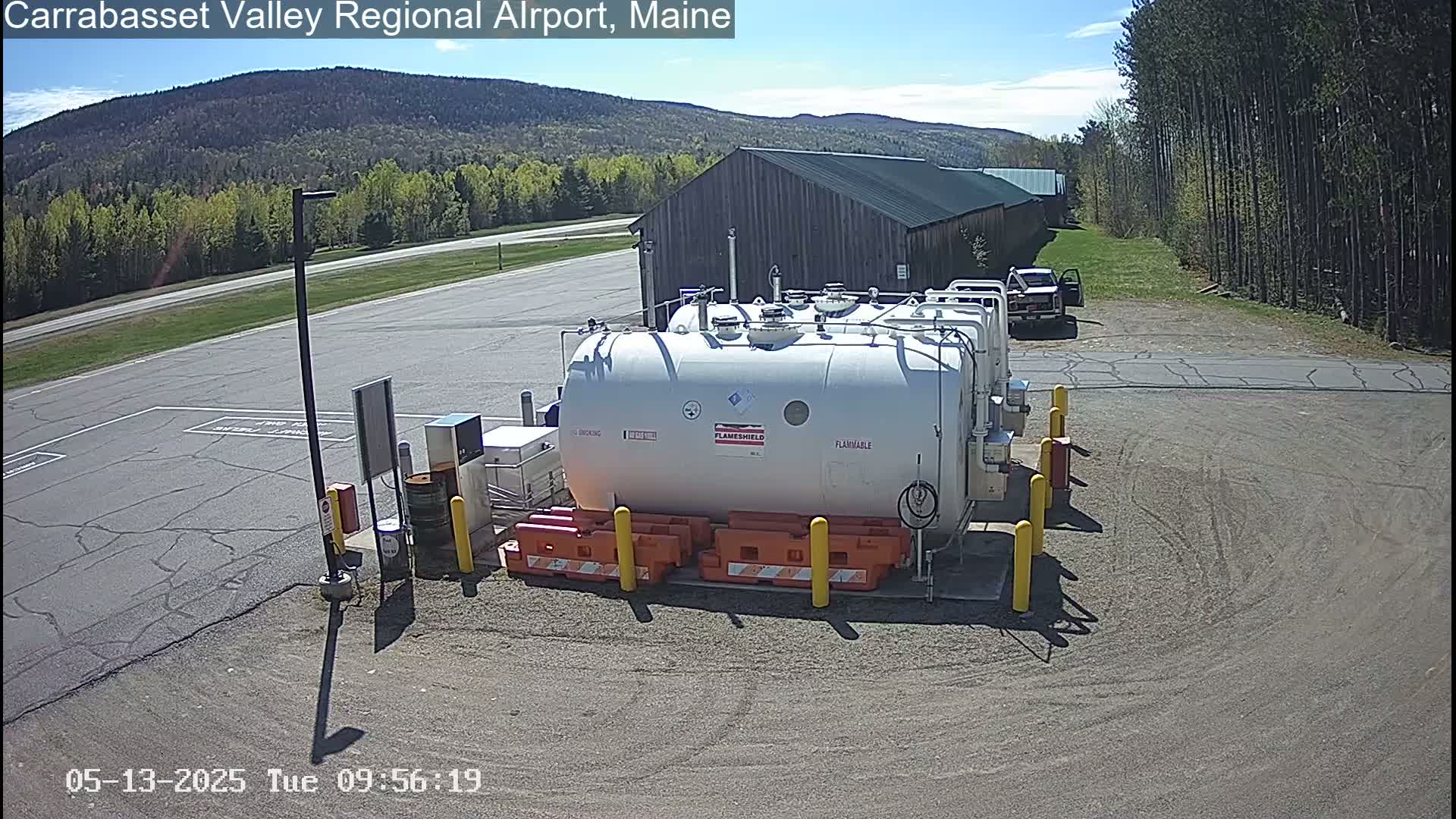 A large white fuel tank sits in a parking lot on a sunny day, near a wood-sided building and a line of trees.