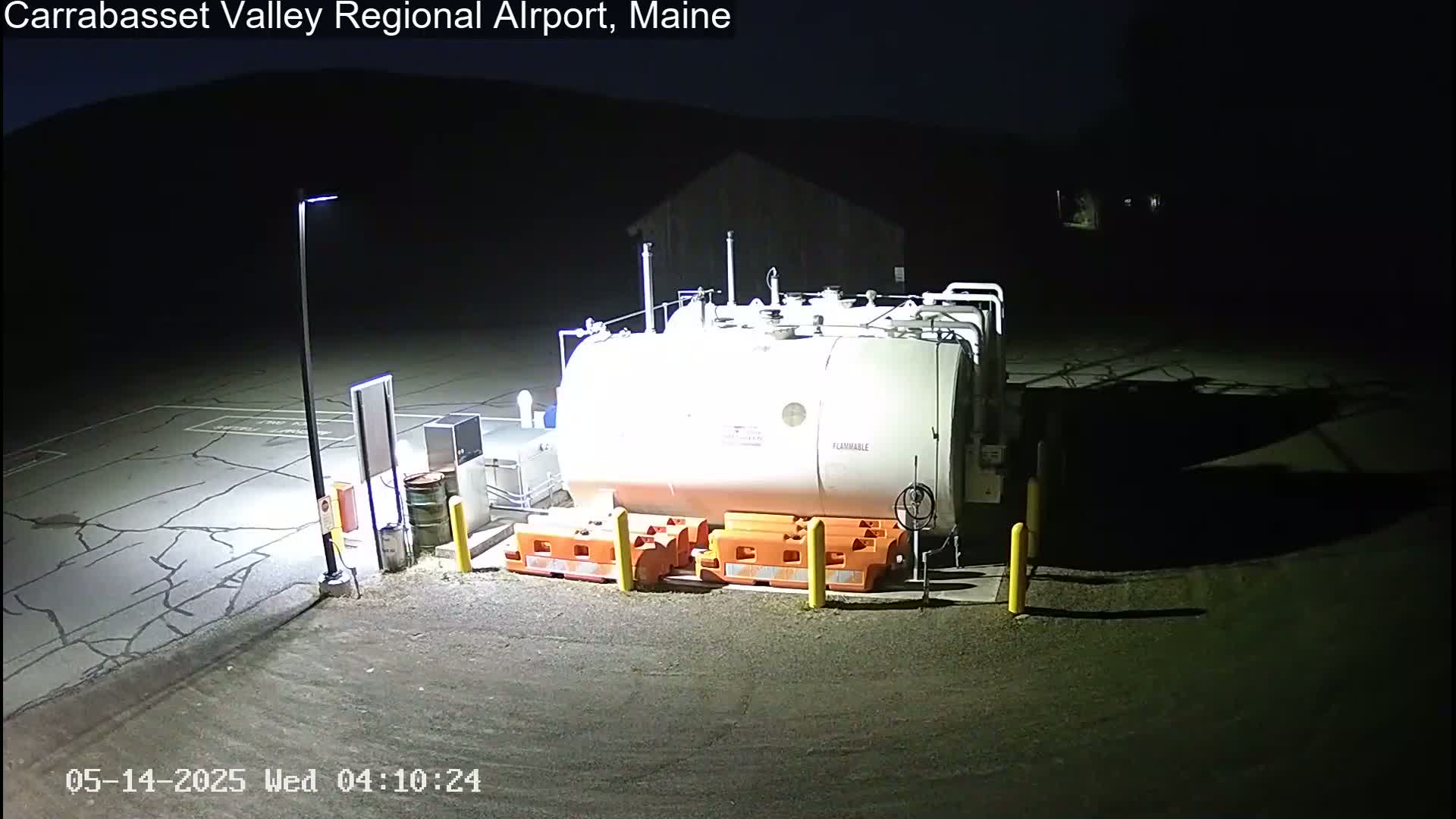 At night, a large white fuel tank sits in a cracked asphalt area, surrounded by orange safety barriers and illuminated by a single light pole.