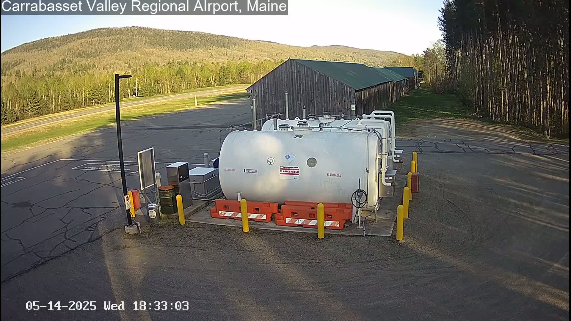 A large white fuel tank sits in a paved area near a long, low wooden building, under a clear sky and surrounded by trees and a grassy field.
