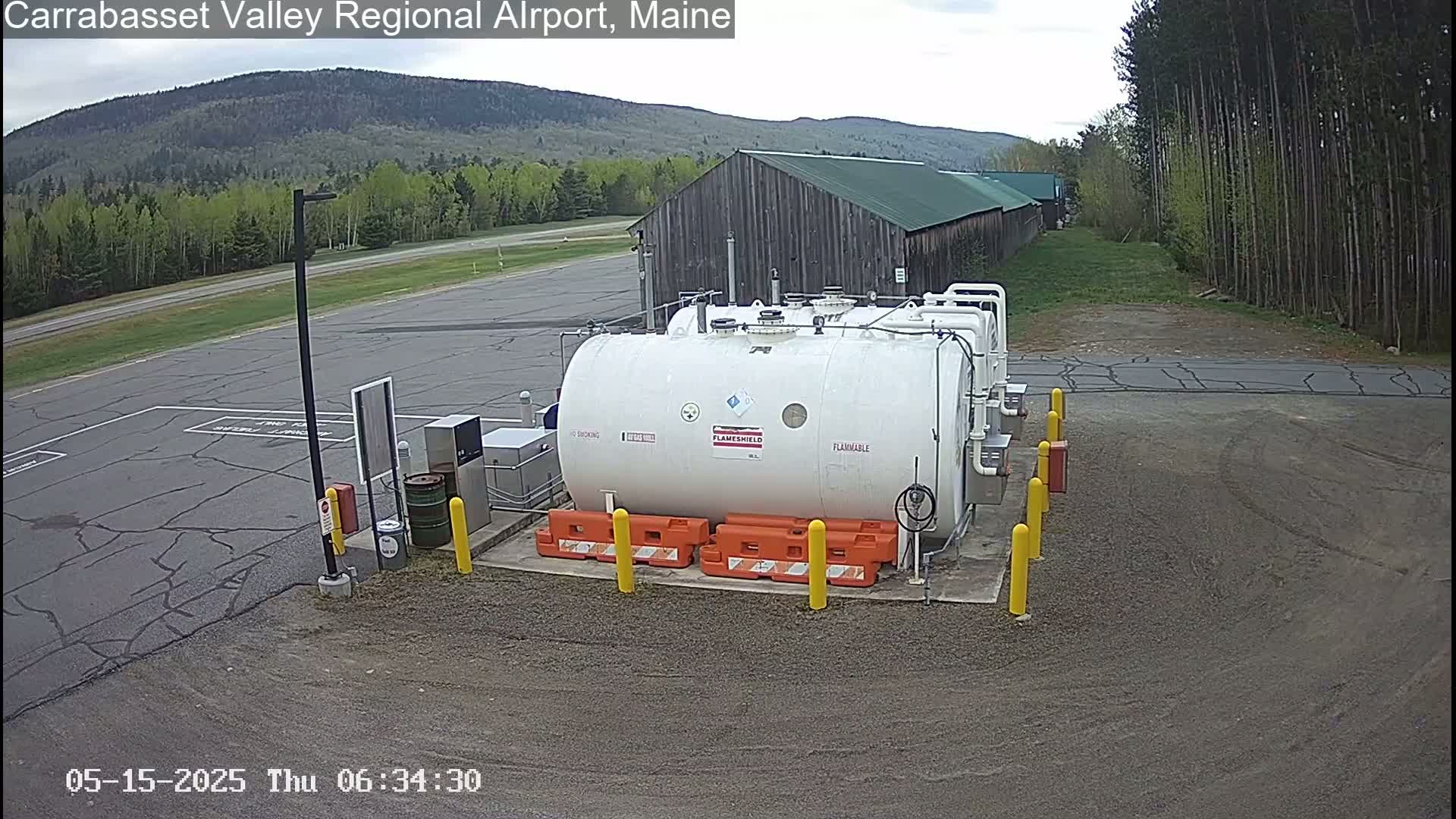 A large white fuel tank sits in a paved area of an airport, surrounded by yellow and orange safety equipment, with a wooden building and a treeline in the background under a mostly cloudy sky.