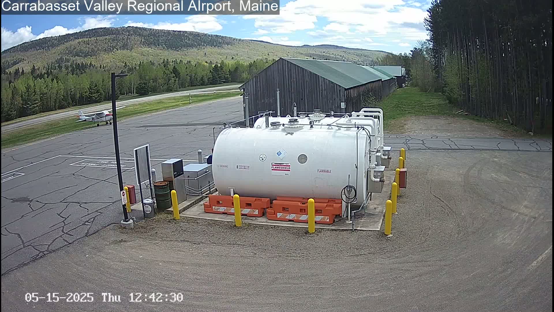 A small airplane sits on a tarmac near a large white fuel tank at an airport on a partly cloudy day, with a wood-sided building and green trees in the background.