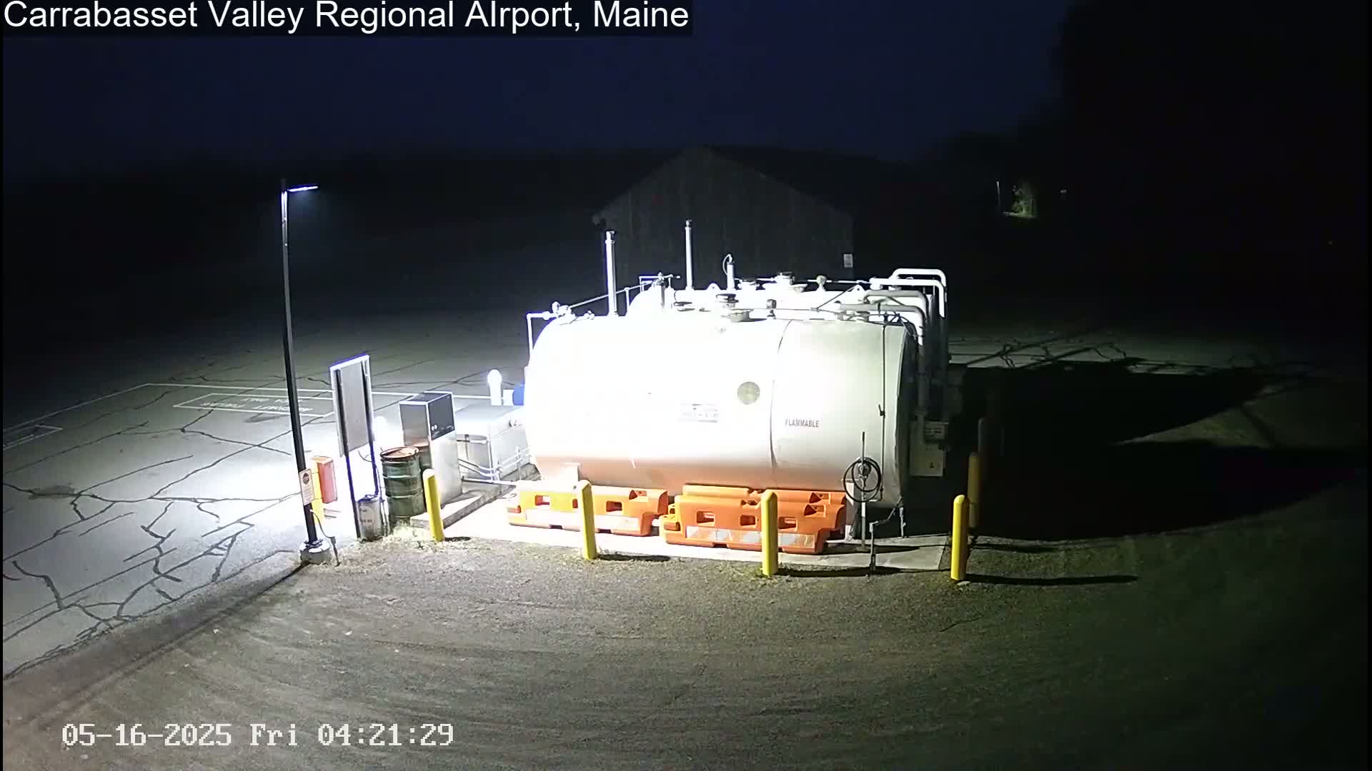 At night, a large white fuel tank sits in a cracked asphalt lot, illuminated by a single lamppost, with orange safety barriers surrounding it.