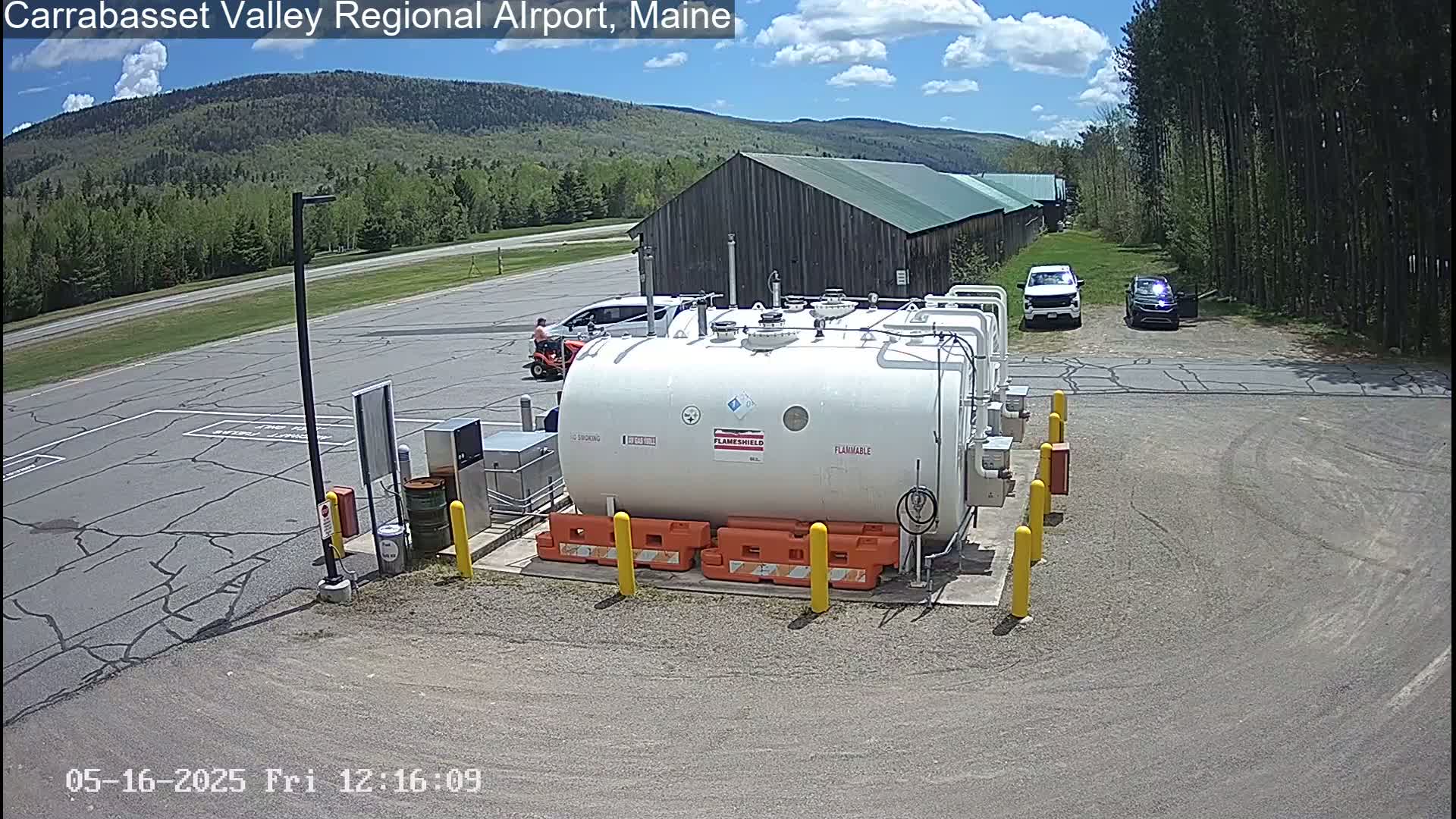 A large white fuel tank sits in a paved area of an airport, near two vehicles and a rustic wooden building, under a partly cloudy sky.