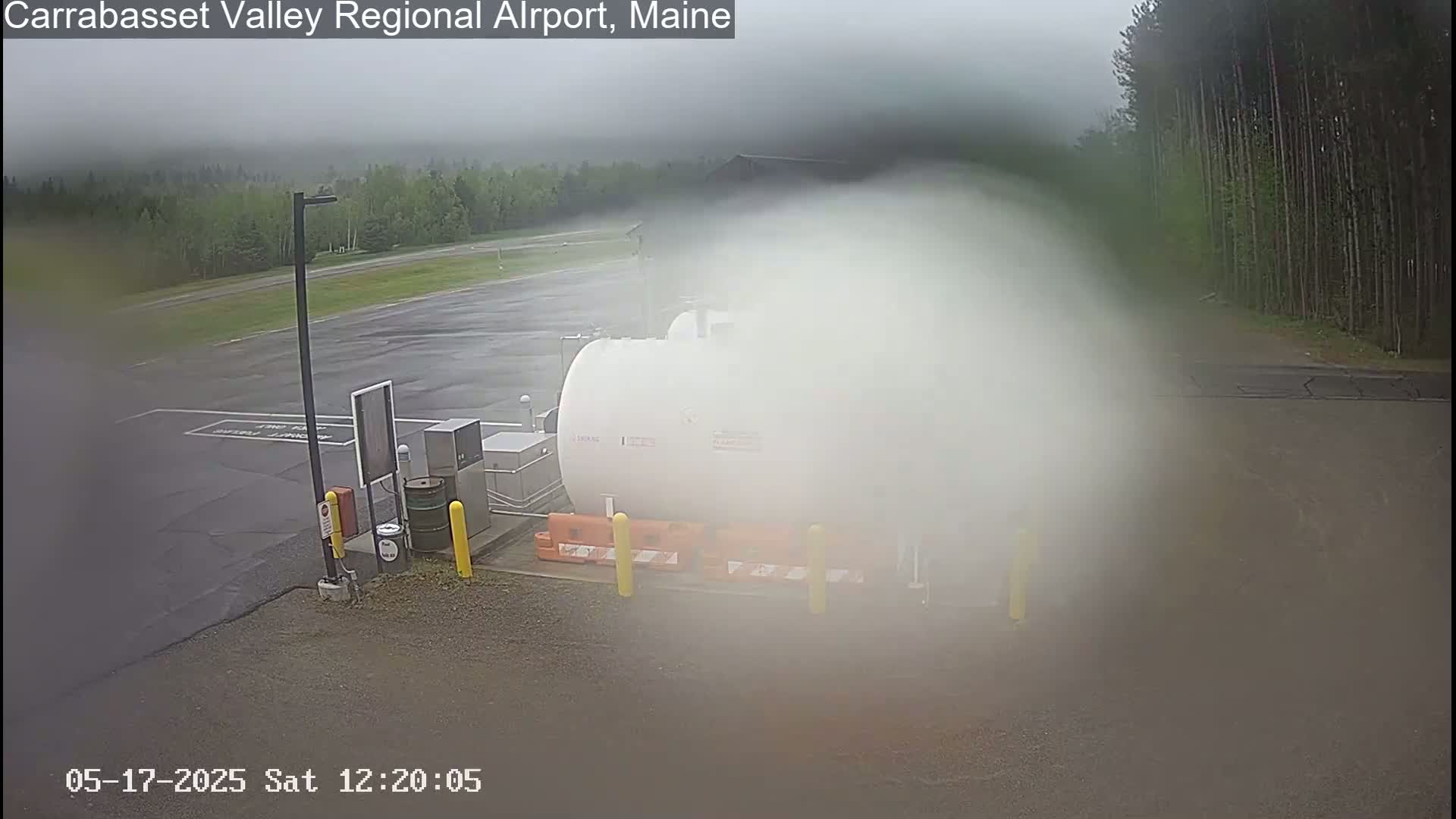 A large white tank sits in a misty, overcast parking lot next to a small airport runway.