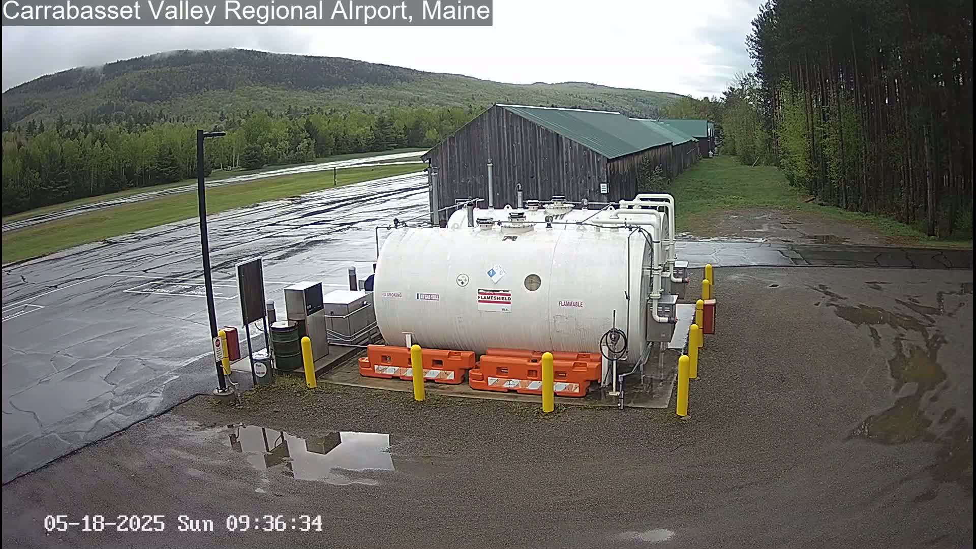 A large white flammable liquid storage tank sits in a wet parking lot next to a long wooden building, with a partly cloudy sky and green forested hills in the background.