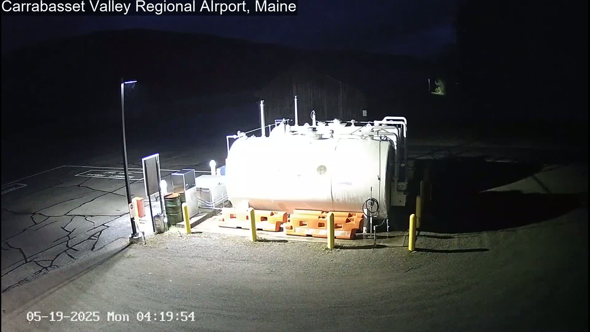 At night, a large, light-colored cylindrical tank sits in a dark, paved area, surrounded by orange safety blocks and illuminated by artificial light.