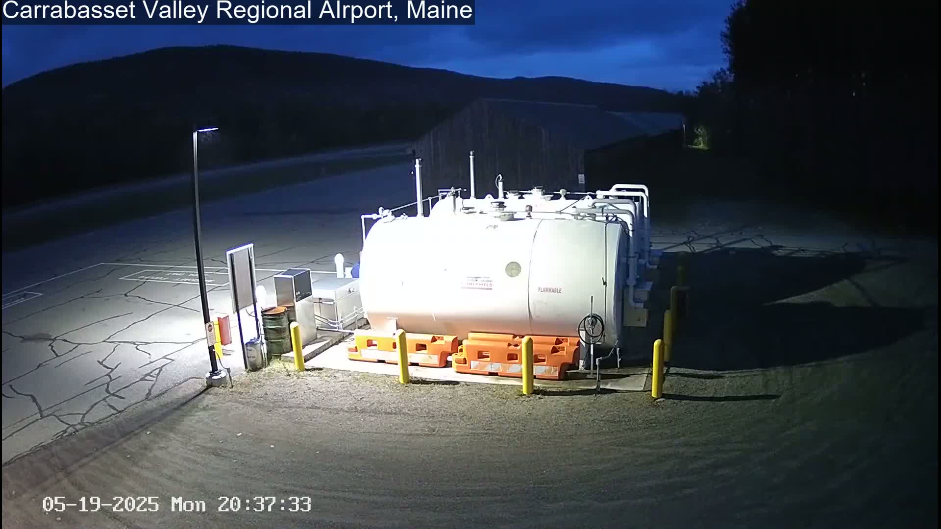 A large, white, cylindrical fuel tank sits in a dark, empty airport parking lot under a night sky.