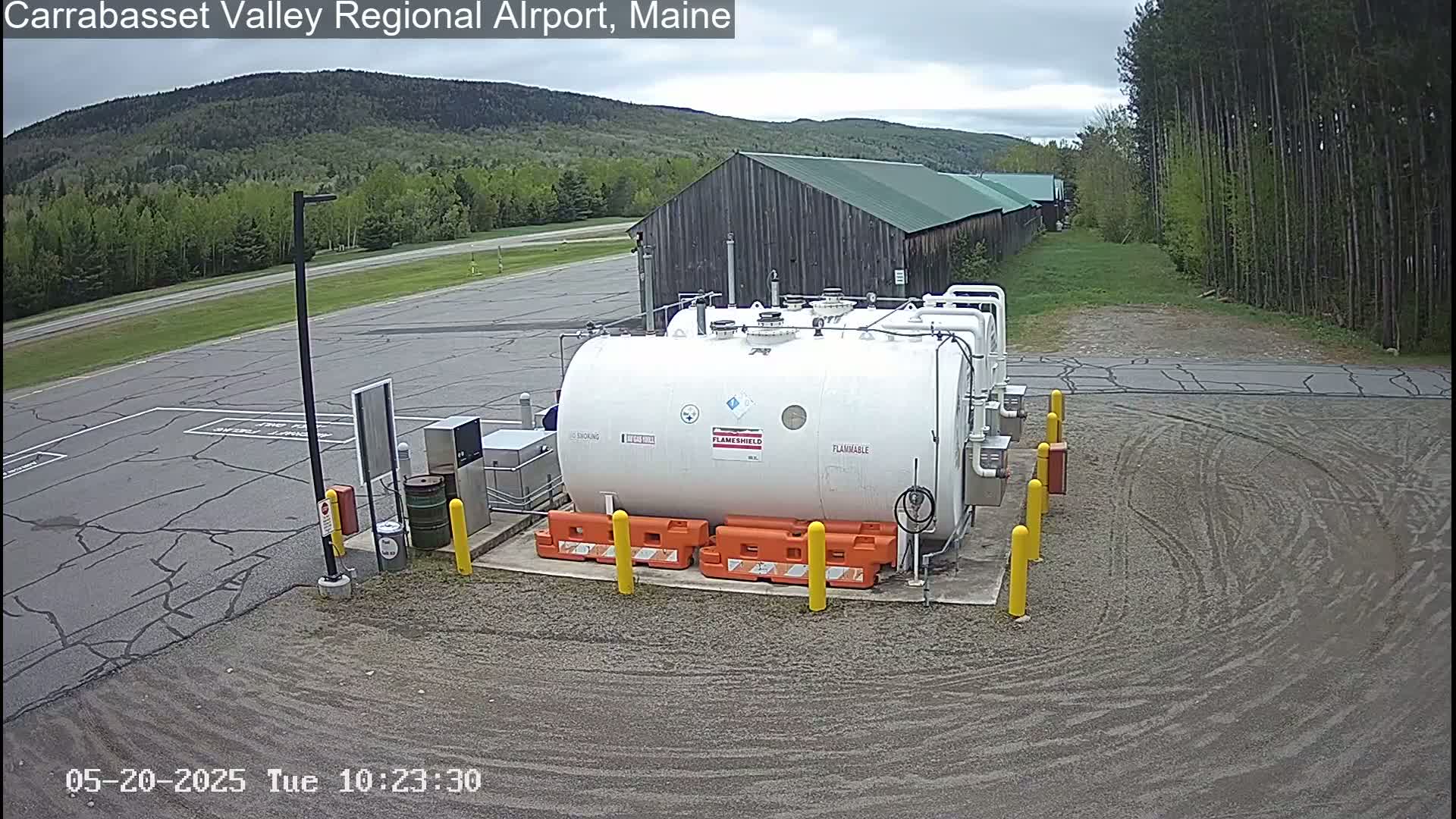 A large white flammable liquid storage tank sits in a gravel area of an airport, with a wood-sided building and forested hills visible in the background under an overcast sky.
