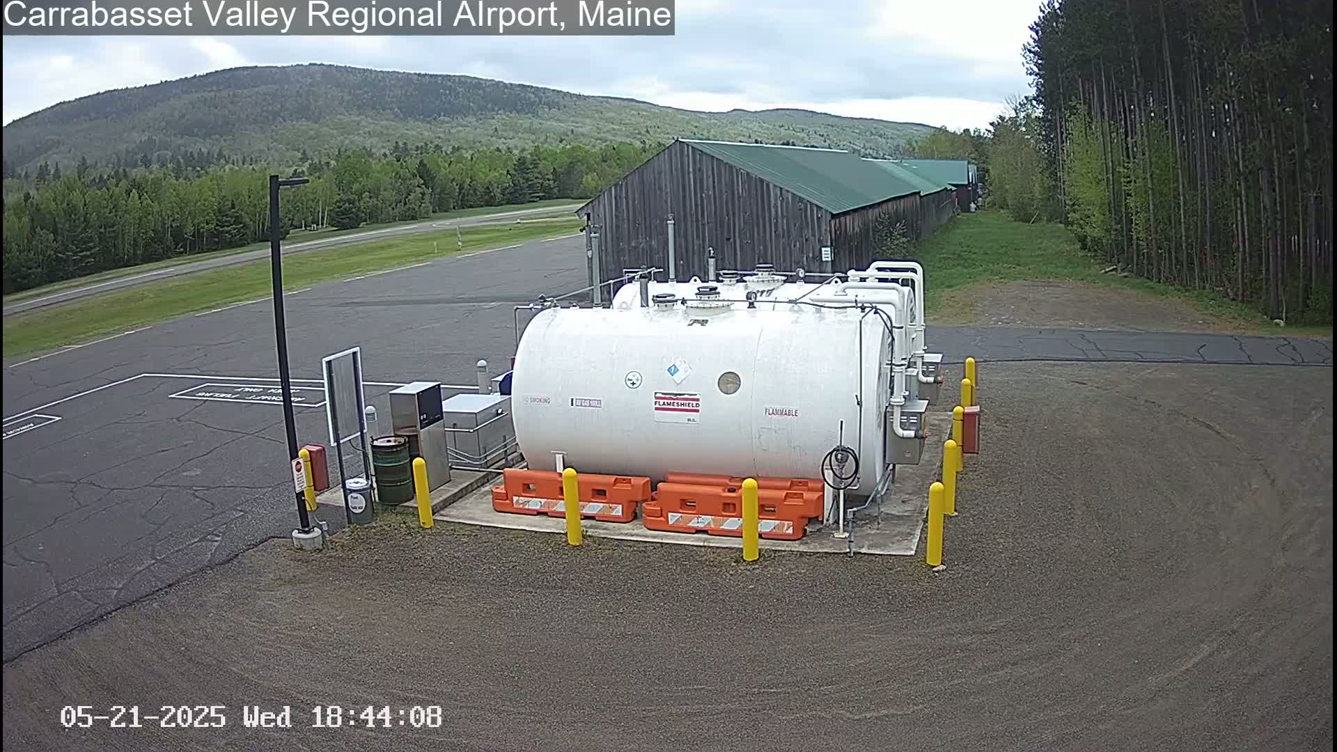 A large white fuel tank sits in a gravel area of an airport, with a wood building and forested hills visible in the background under an overcast sky.