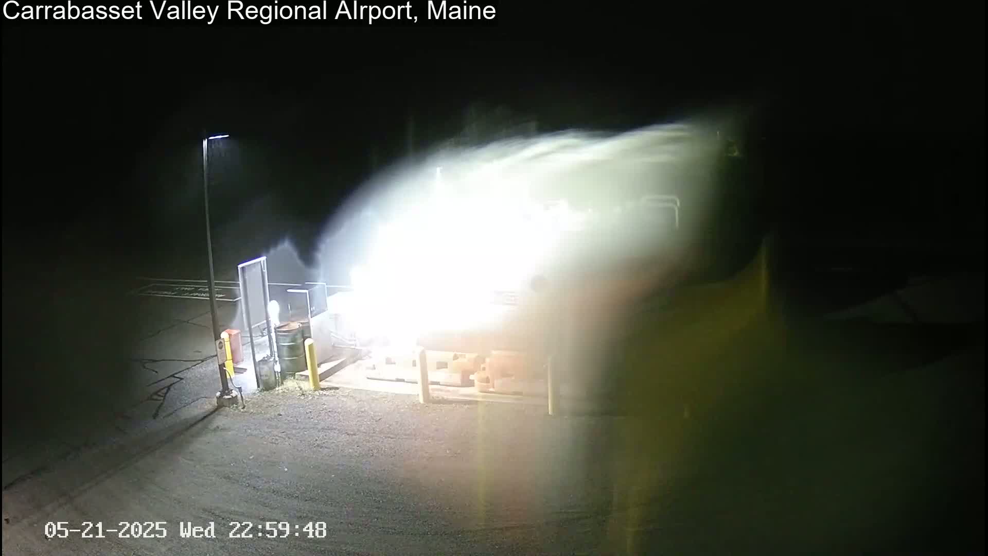 A nighttime scene shows a bright, blurry area, possibly from snow or blowing dust, near a building and a light pole in an otherwise dark parking lot.
