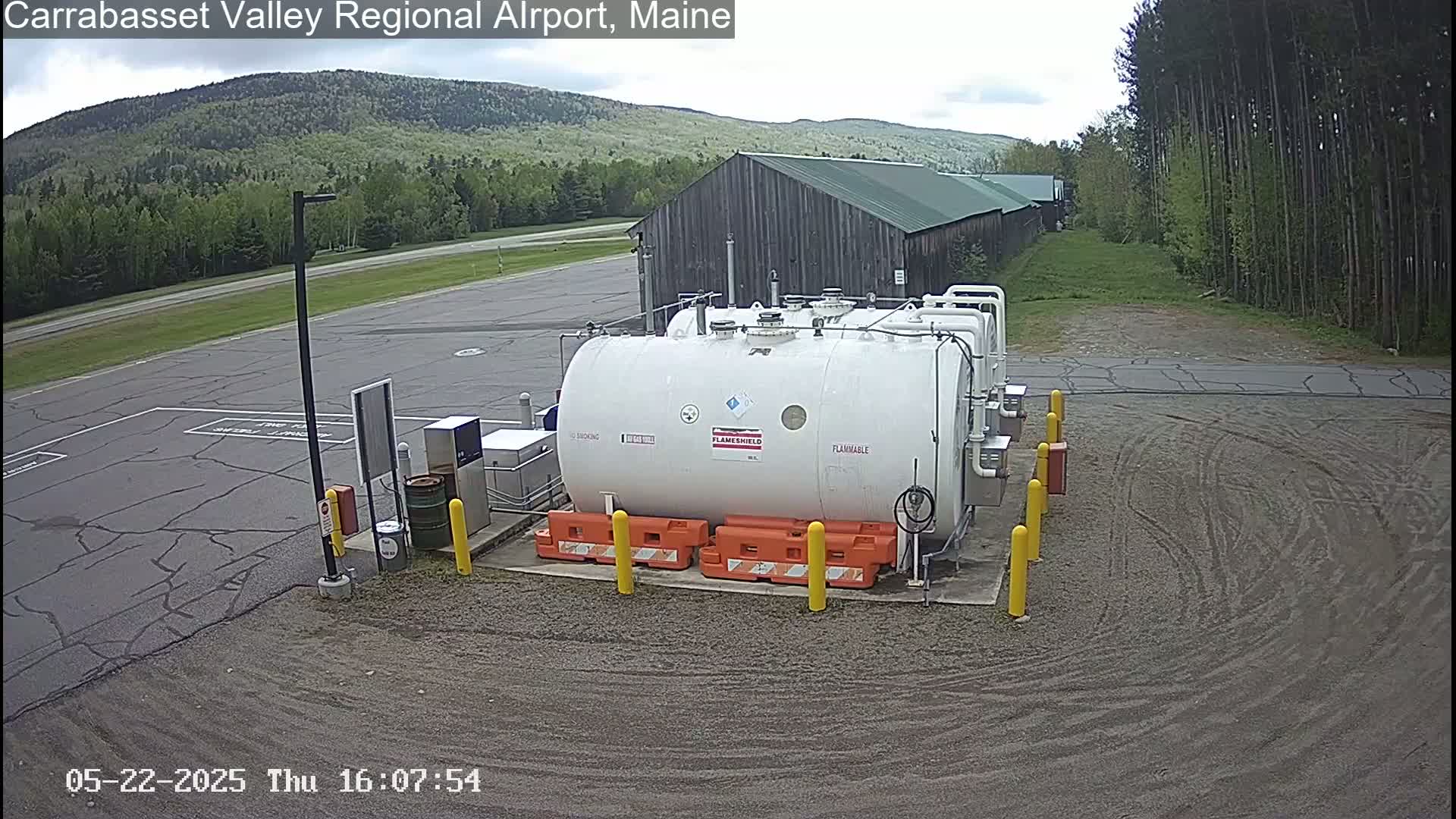 A large white fuel tank sits in a gravel area of an airport, with a wood building and green forested hills visible in the background under a mostly cloudy sky.