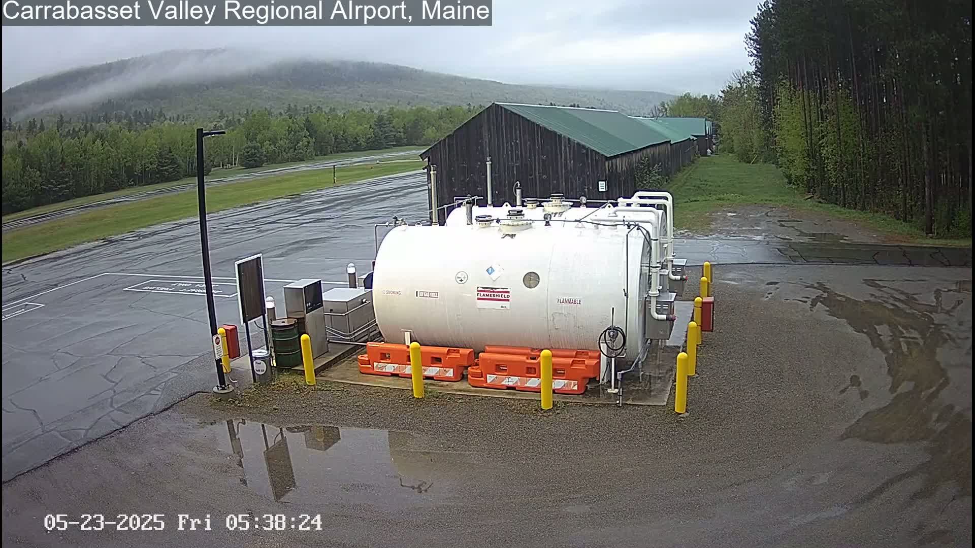 A large white flammable liquid storage tank sits in a wet asphalt area next to a dark-colored building, with a misty mountain visible in the background under an overcast sky.