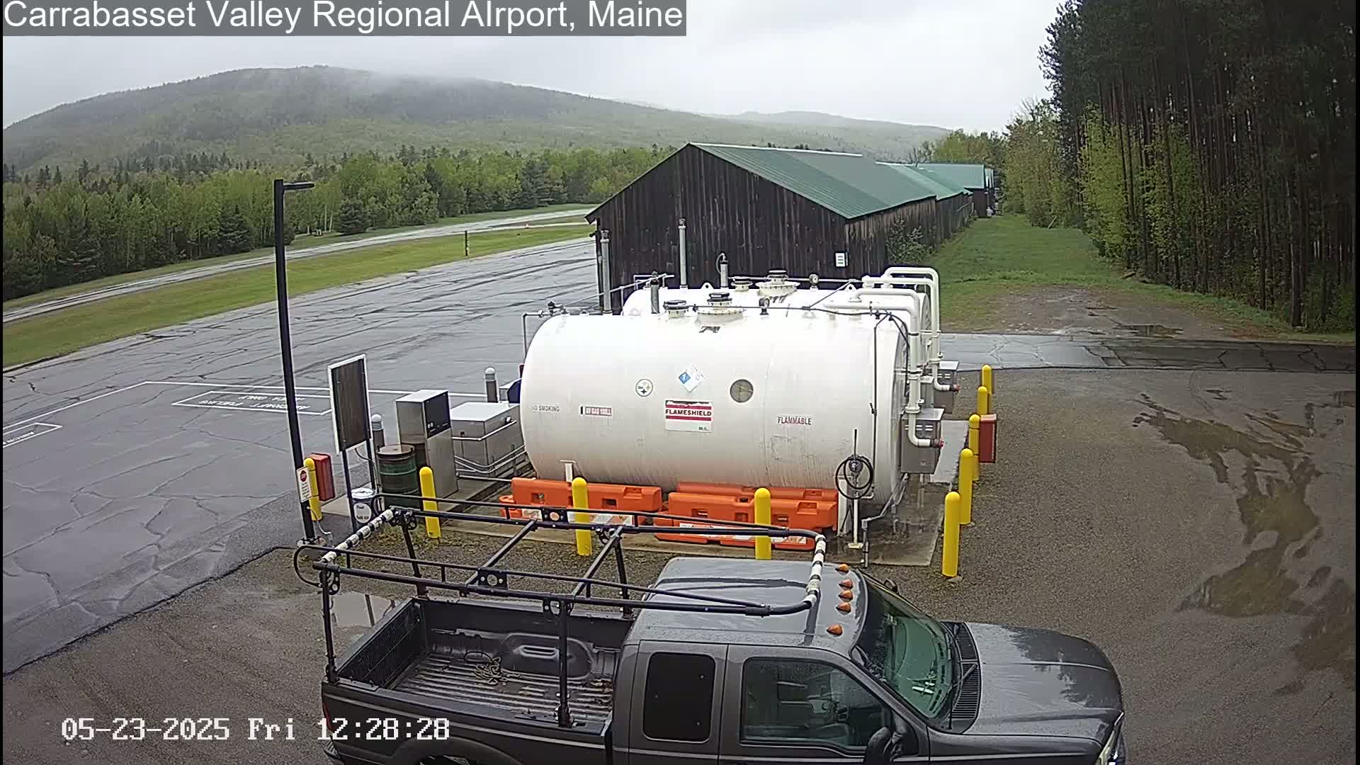 A large white cylindrical tank sits in a wet, paved area next to a dark-colored pickup truck, with a dark-colored building and a line of trees in the background under an overcast sky.