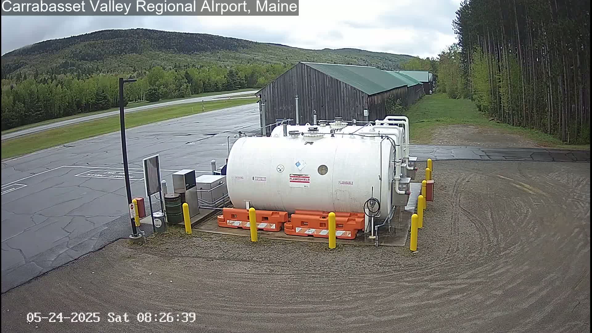 A large white cylindrical tank sits in a paved area of an airport, with a long building and green, forested hills visible in the background under an overcast sky.
