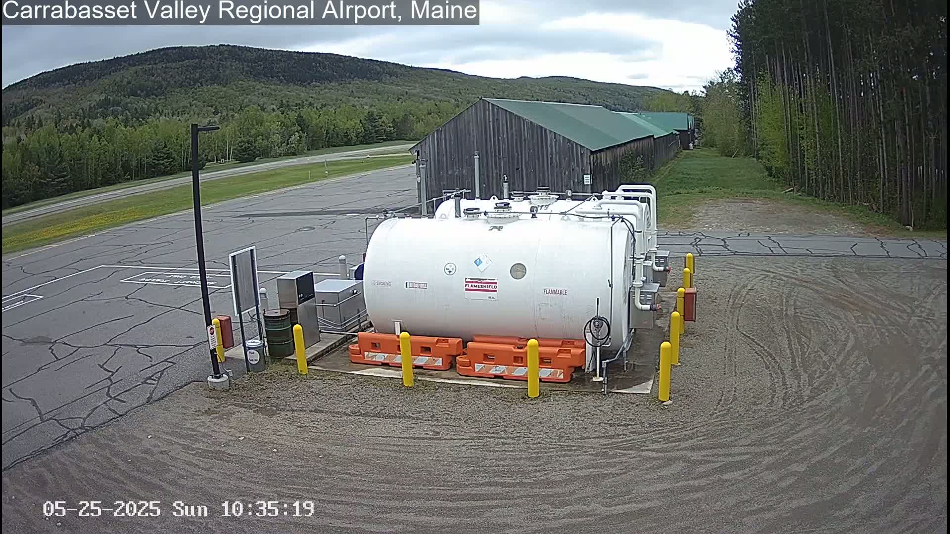 A large white fuel tank sits in a gravel area of an airport, with a gray overcast sky and green trees and a brown building in the background.