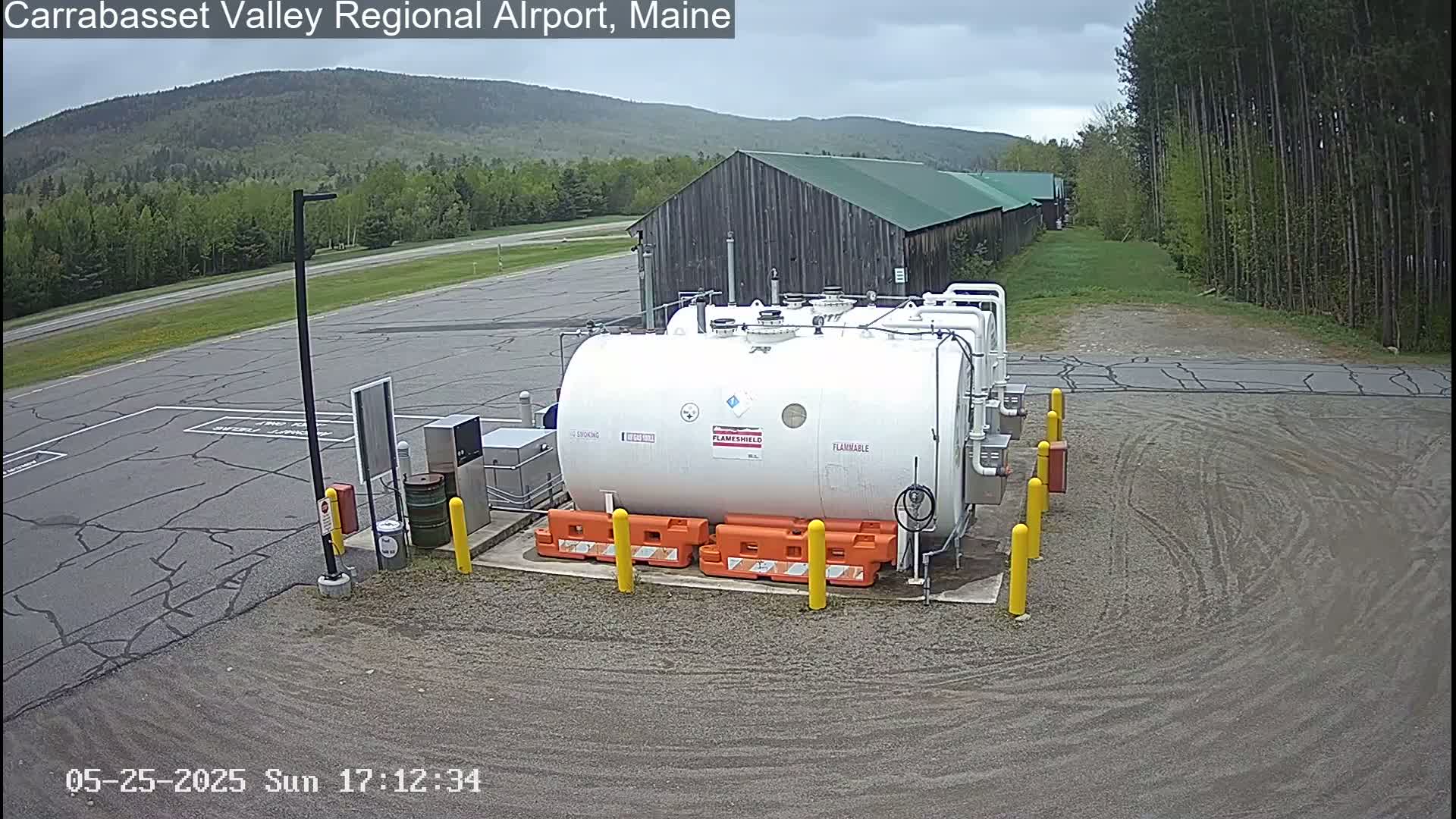 A large white fuel tank sits in a gravel area of an airport, with a wood building and green trees in the background under an overcast sky.
