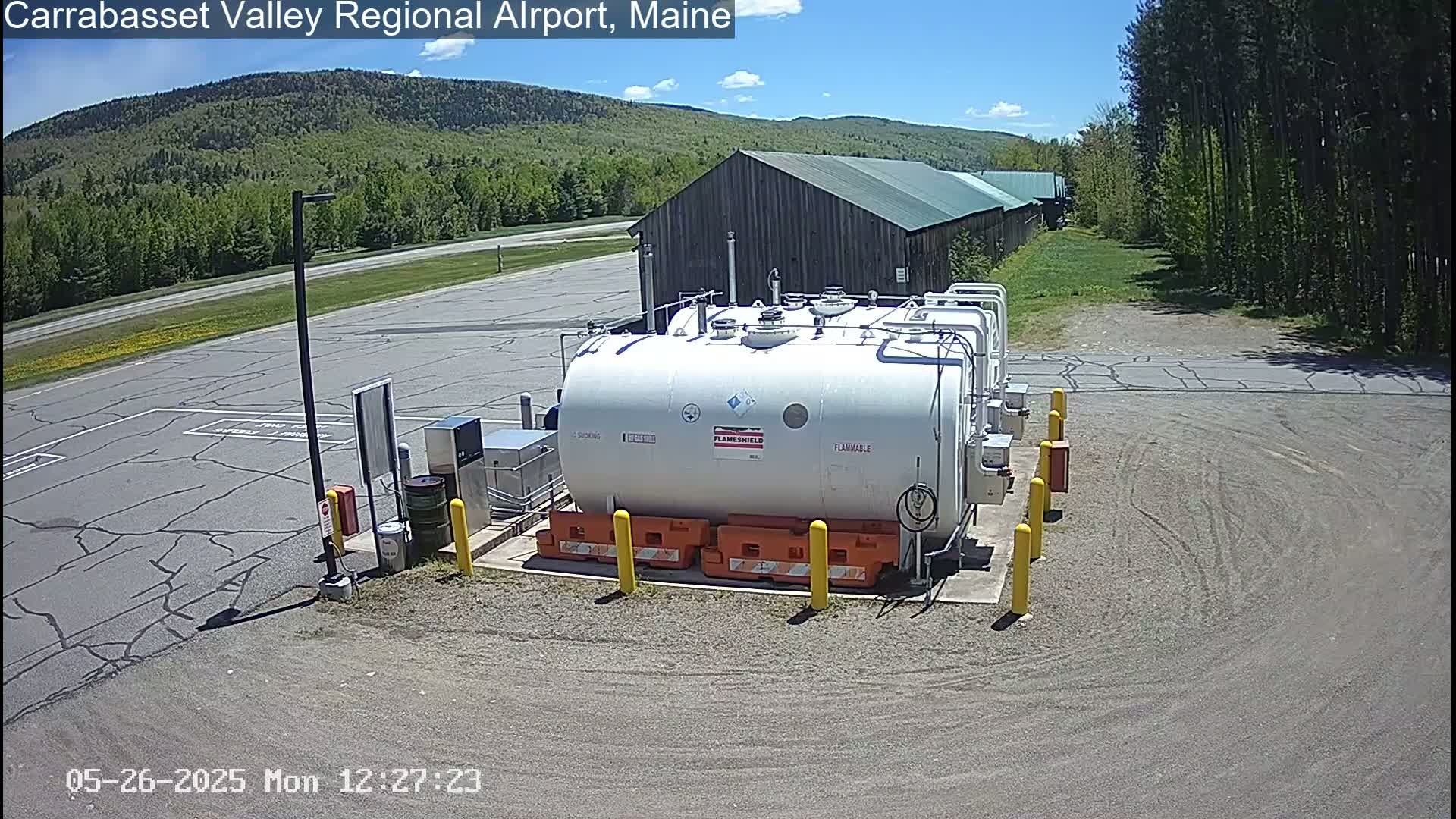 A large, white, flammable liquid storage tank sits in a paved area of an airport on a sunny day, with a rustic building and a forested hillside in the background.