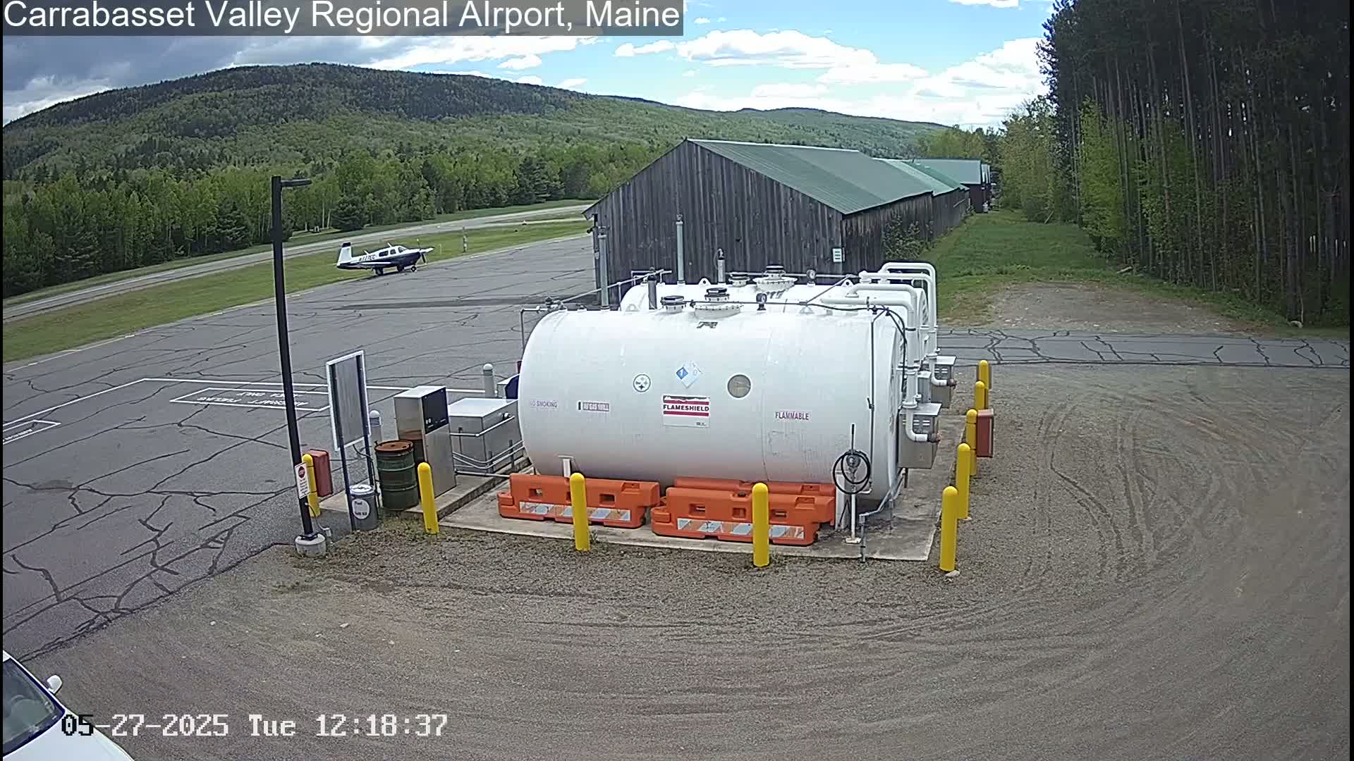 A large white fuel tank sits in an airport parking lot under a partly cloudy sky, with a small airplane and a wooded hillside in the background.