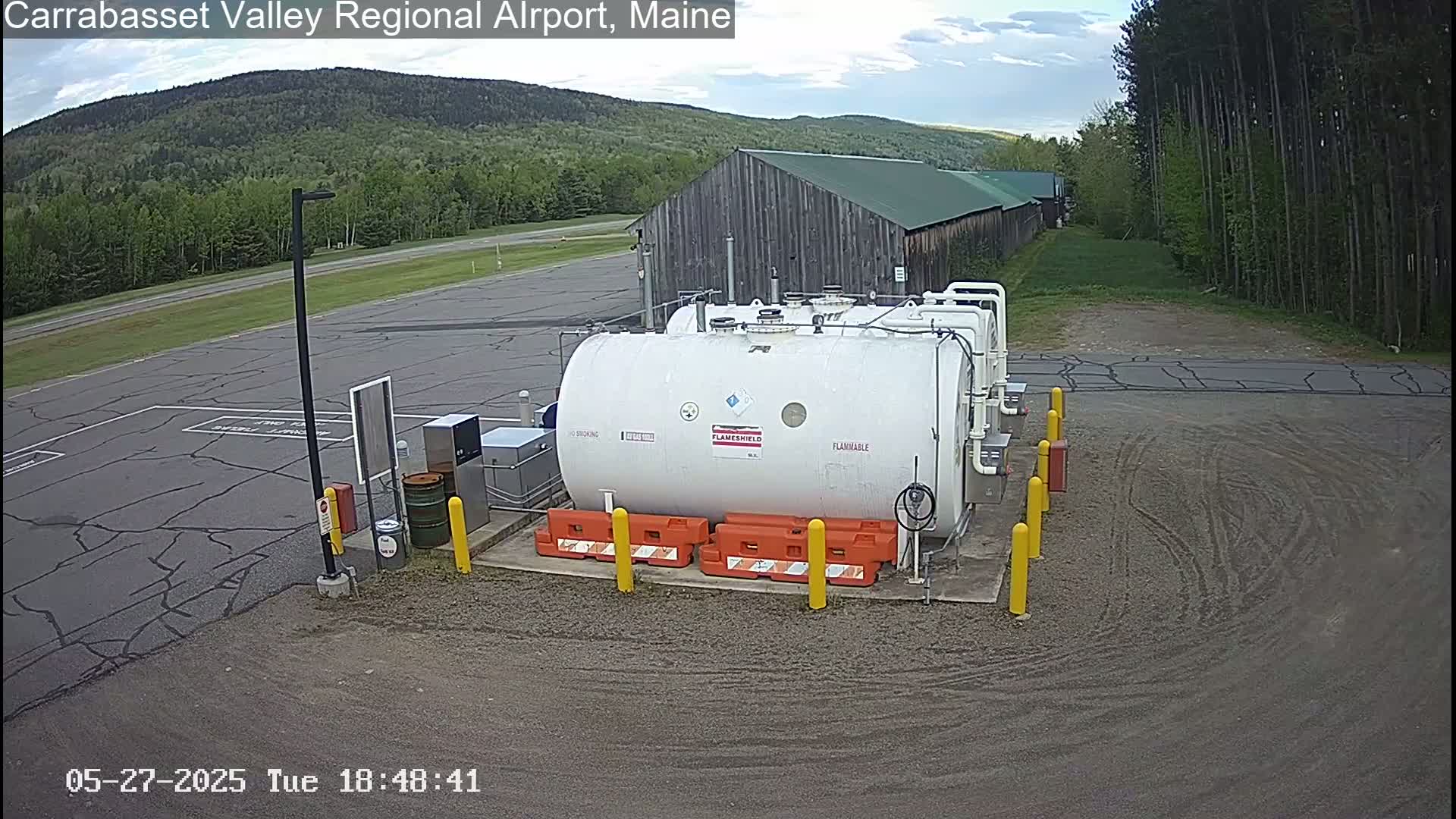 A large white fuel tank sits in a paved area next to a rustic wooden building, under a partly cloudy sky.