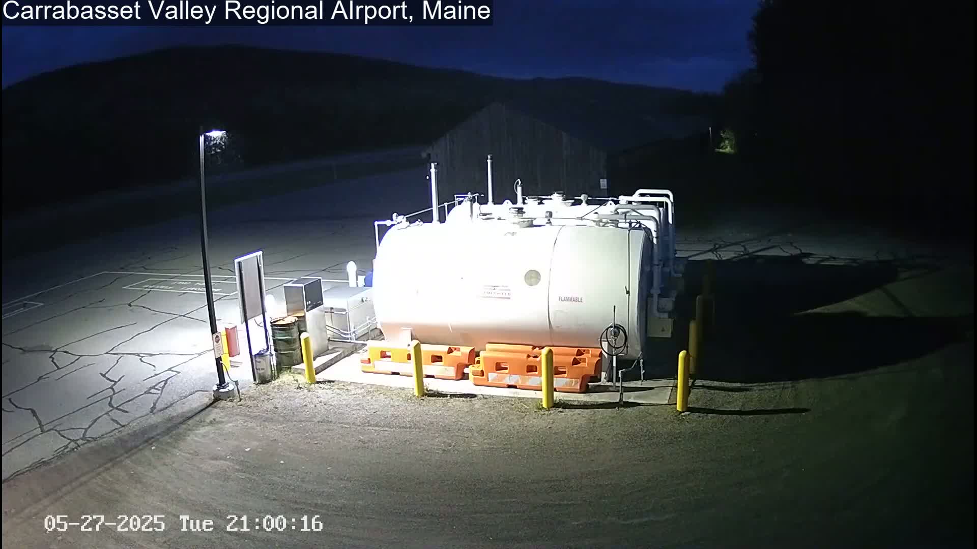 A large, white, cylindrical fuel tank sits in a dark, cracked asphalt lot at night, illuminated by a single lamppost.
