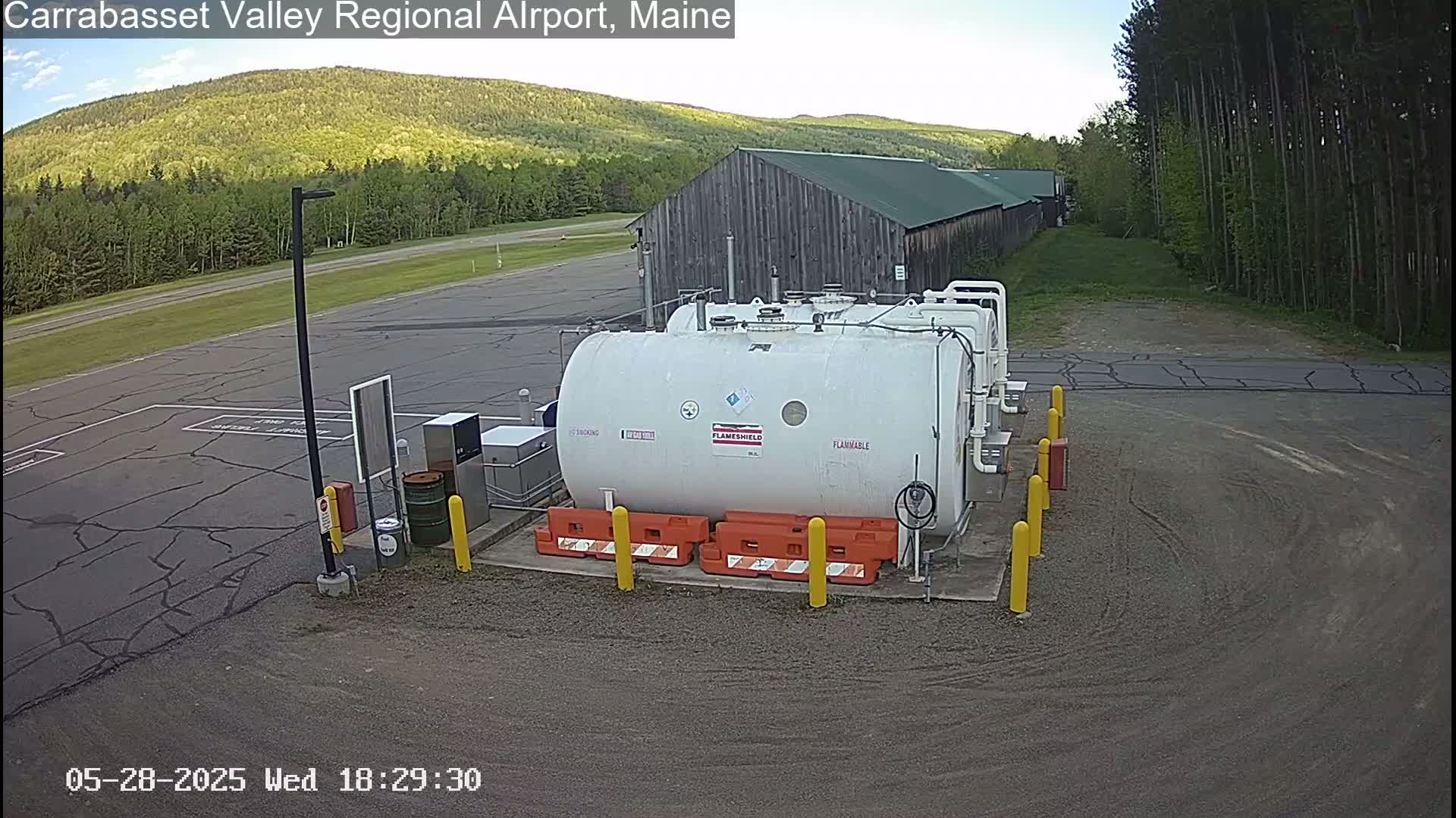 A large white fuel tank sits in a paved area of an airport, under a mostly clear sky, with a wooded hillside and a long building in the background.