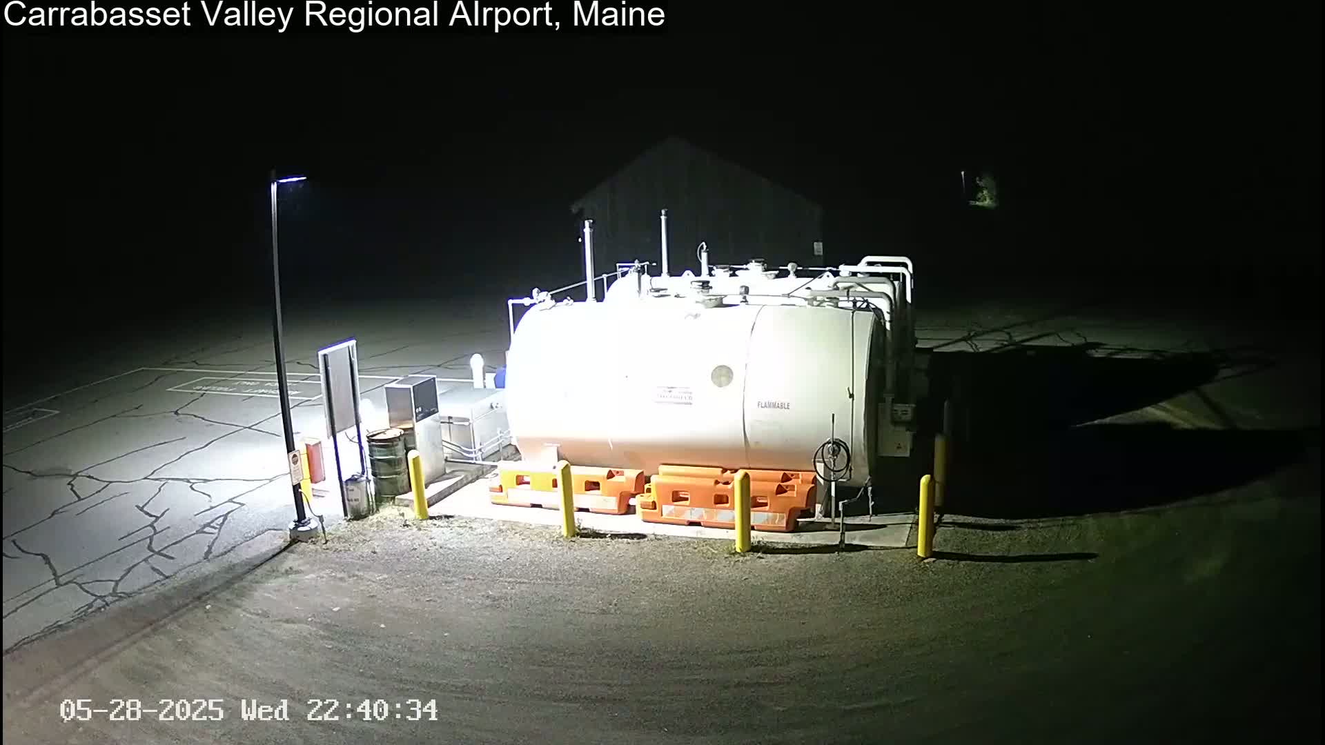 At night, a large, white, cylindrical fuel tank sits in a cracked asphalt area, illuminated by a single light pole.