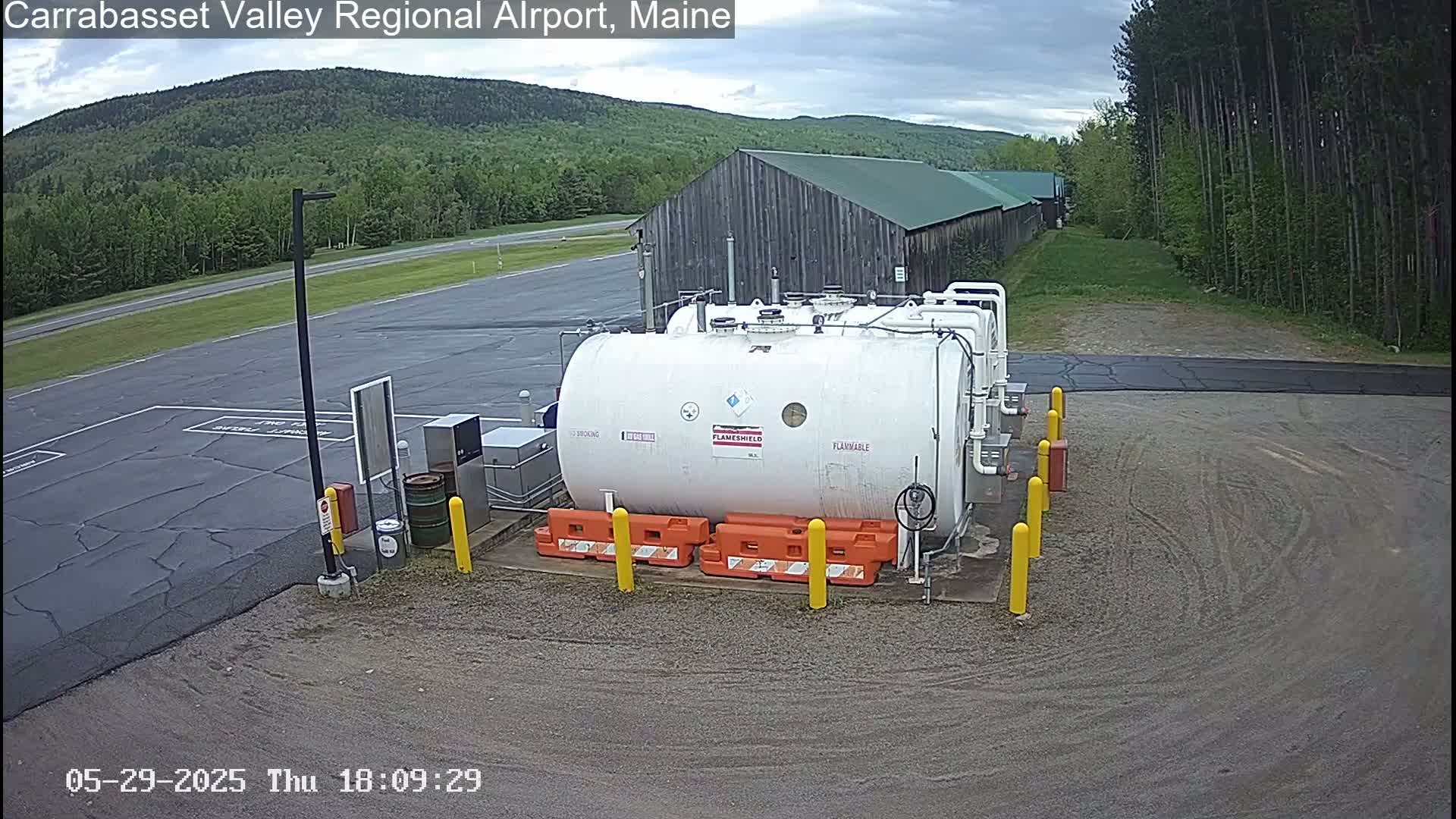 A large white flammable liquid storage tank sits in a paved area of an airport, with a rustic wooden building and green forested hills visible in the background under a cloudy sky.