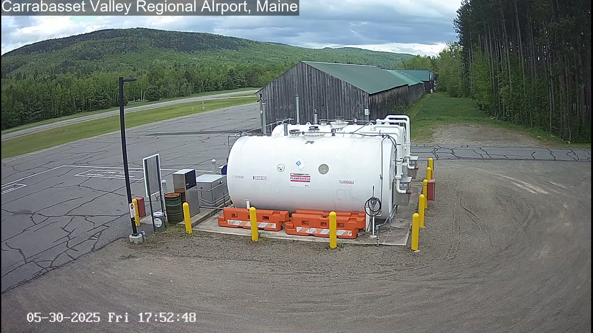 A large white flammable liquid storage tank sits in a gravel parking lot next to a long wooden building, under a partly cloudy sky.