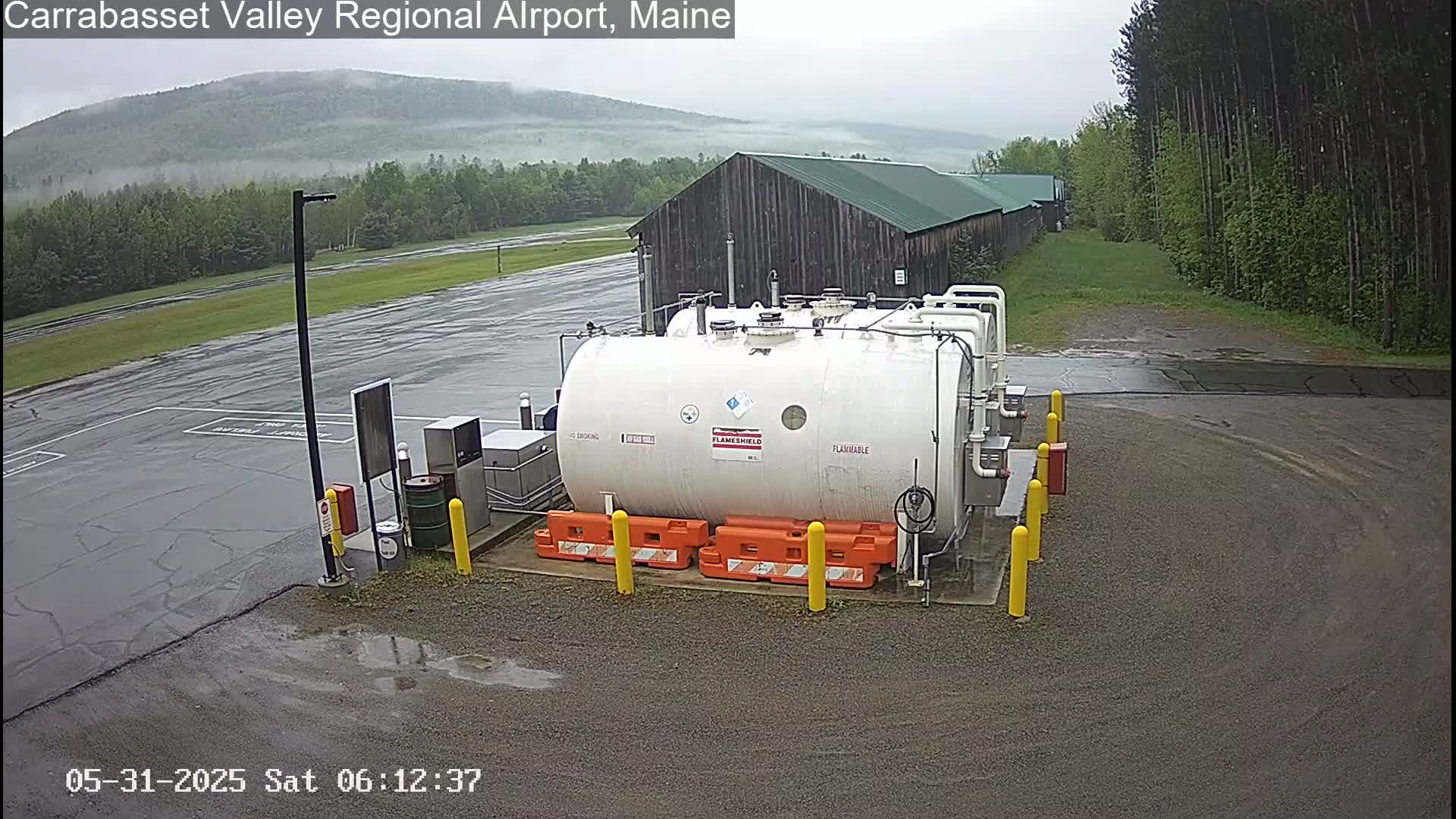 A large white cylindrical tank sits in a wet, paved area near a wooden building, with a misty mountain range visible in the background under an overcast sky.