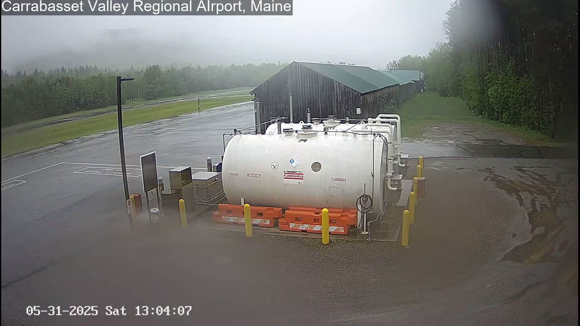 A large white flammable liquid storage tank sits in a wet airport parking lot next to a dark-colored building, under a rainy sky.